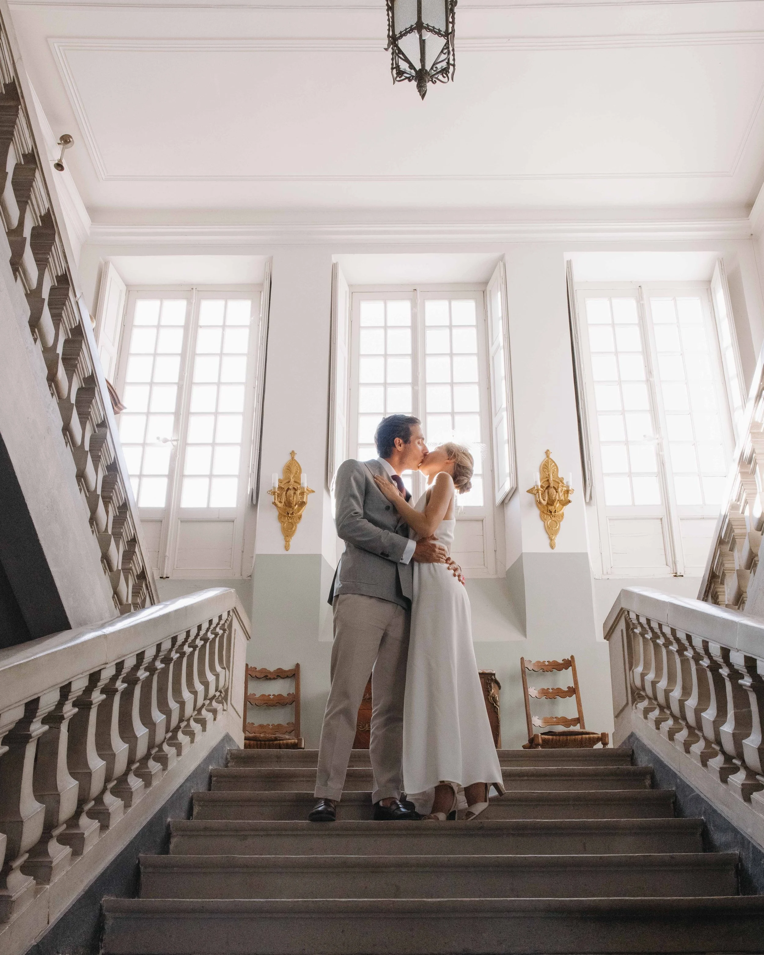 Un couple en costume de mariage s'embrasse au sommet d'un escalier dans une grande salle lumineuse avec de hauts plafonds et de grandes fenêtres. Mariage au chateau de lachal dans le rhone alpes à côté de Lyon. Cérémonie civil et laïque. 