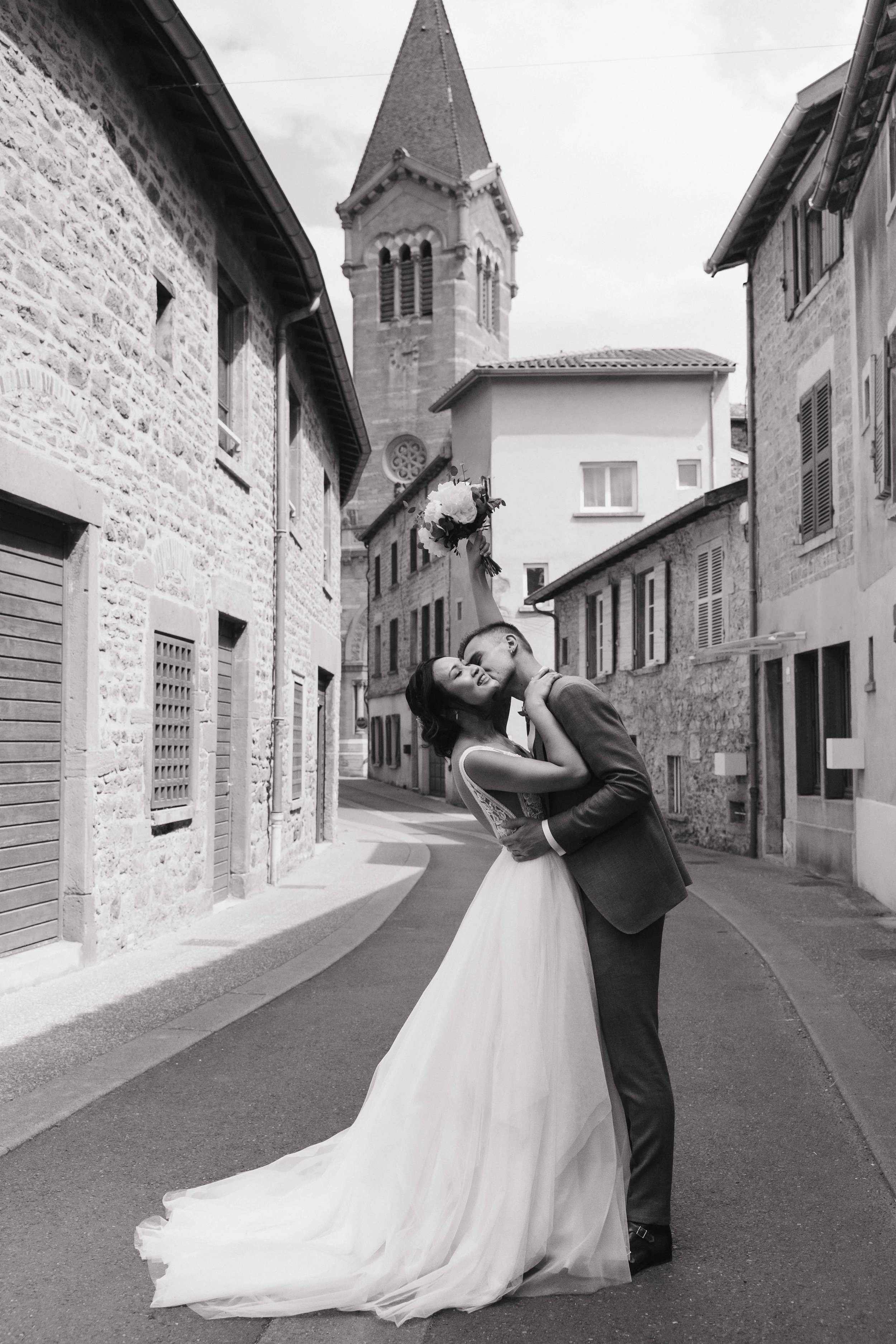 Un couple de mariés qui s'embrassent dans une rue pavée, avec un bâtiment en pierre et une église en arrière-plan, en noir et blanc. Dans le beaujolais à Denicé, village en pierre dorées