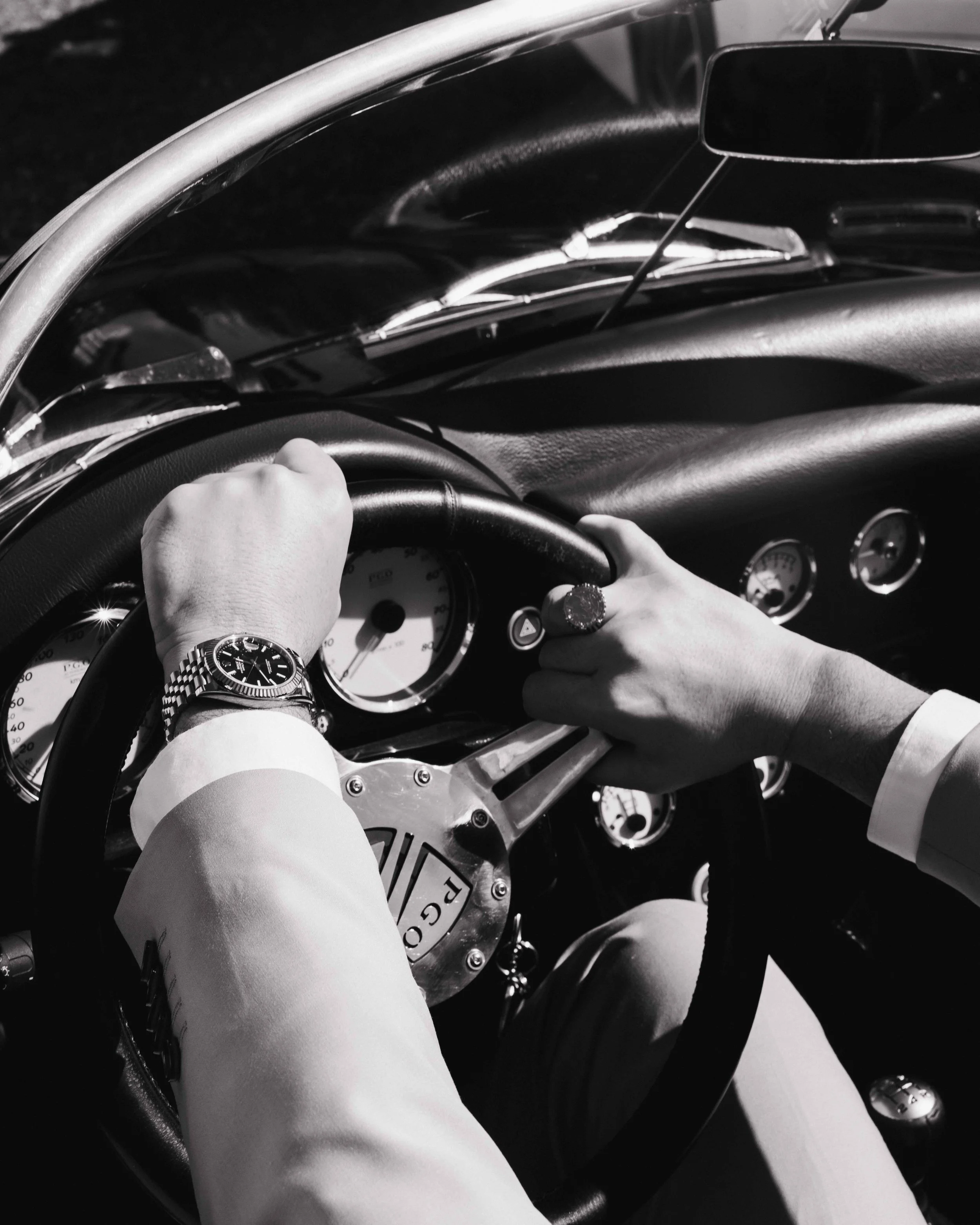 Deux mains sur le volant d'une voiture classique ancienne en noir et blanc, avec un cadran de montre bracelet en acier au poignet gauche. Photographe mariage Isère, ferme du rocher, cérémonie civil et laïque. Bordeaux et Lyon et destination wedding. 