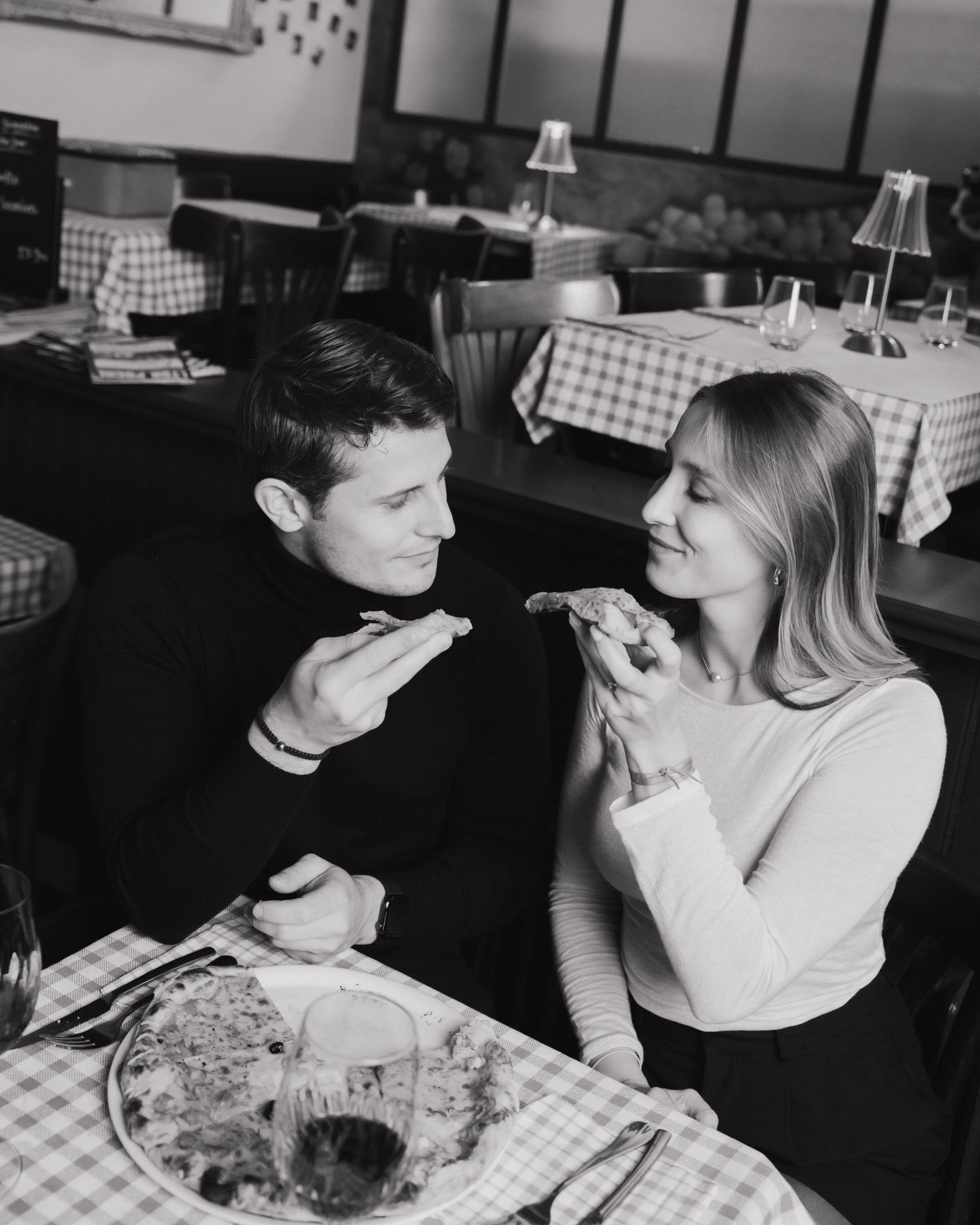 Un couple partage une pizza dans un restaurant, la femme tend une part de pizza à l'homme, tous deux sourient. session engagement couple Lyon 6eme, brotteaux, photographe mariage bordeaux et Lyon et destination wedding