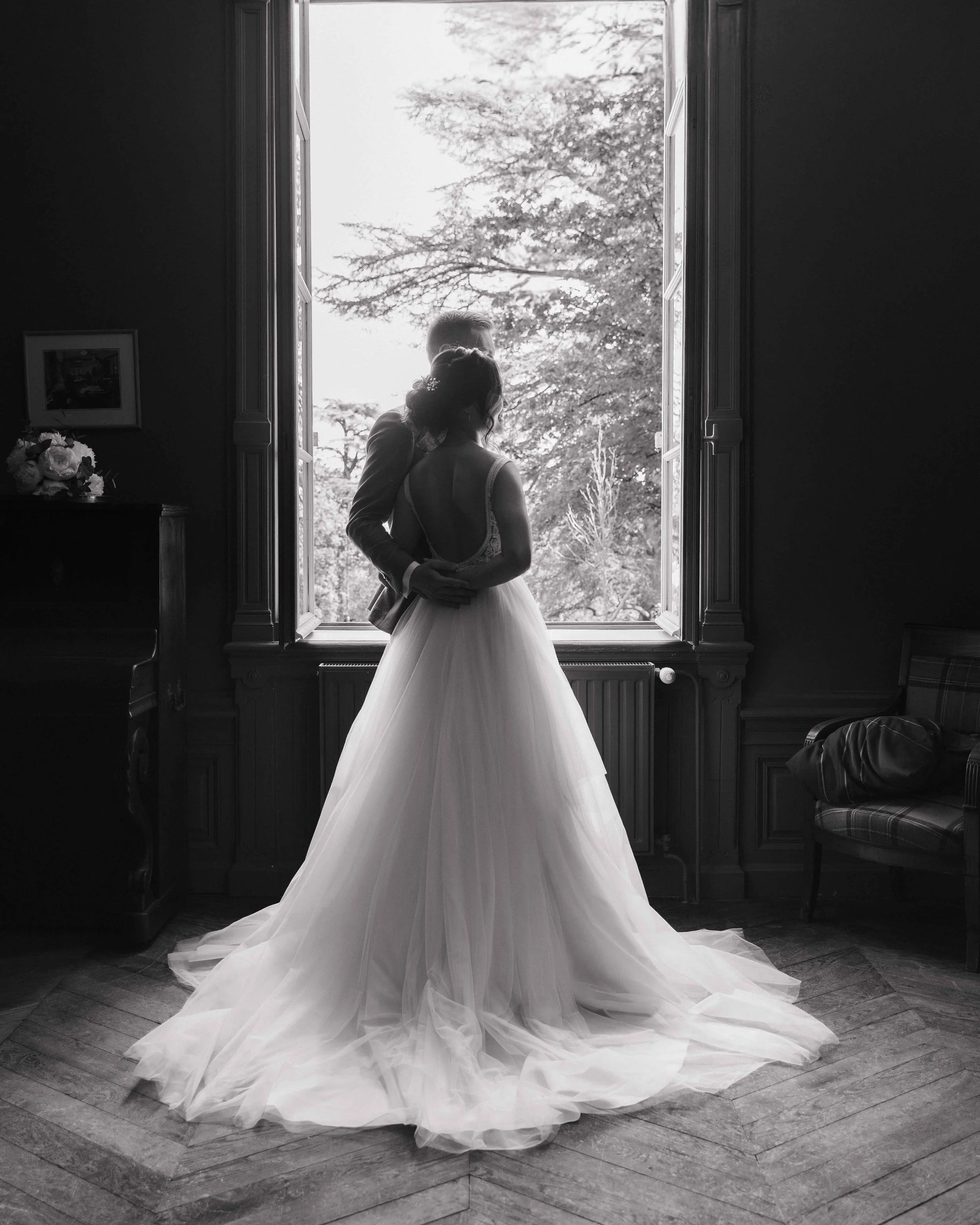 Une mariée en robe de mariage blanche se tenant près d'une fenêtre ouverte, avec un paysage d'arbres en arrière-plan, dans une pièce sombre. Domaine de tourieux à Savigny dans le beaujolais à Lyon. Mariage cérémonie laïque, photographe Lyon