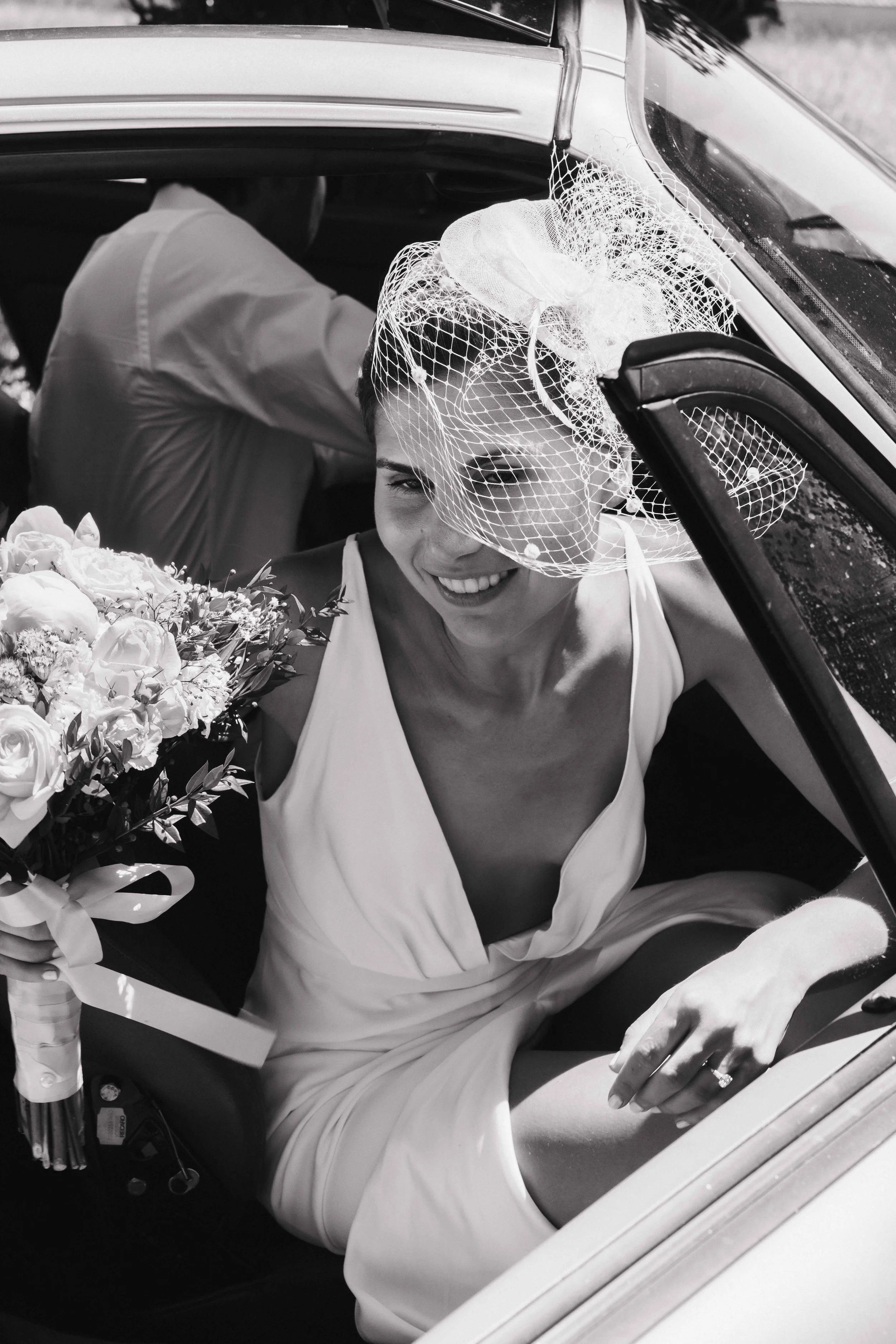 Une femme en robe de mariée souriante, portant un voile, tenant un bouquet de fleurs, assise dans une voiture. Mariage au chateau de lachal dans le rhone alpes à côté de Lyon. Cérémonie civil et laïque. Robe de mariées et costume homme. Photographe 