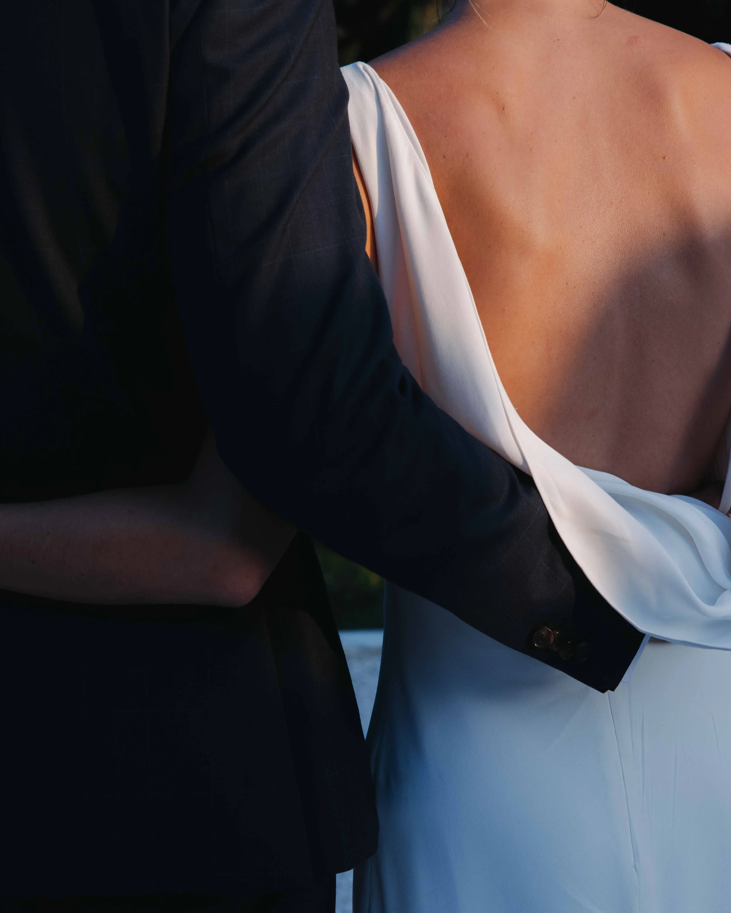 Une personne en robe blanche avec décolleté profond et un homme en costume noir l'encerclent, lors d'une séance photo ou un évènement spécial. Domaine du bijoutier à grignan, drome provençal, coucher de soleil, session couple, mariage religieux 