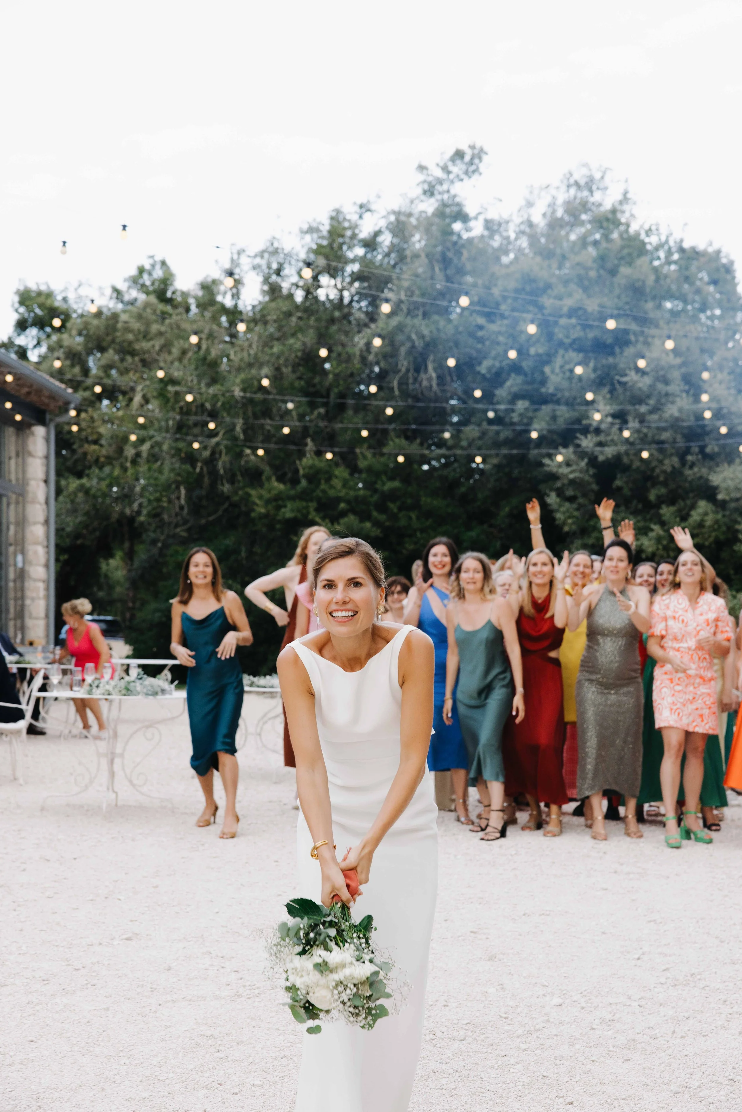 Une mariée souriante en robe blanche tenant un bouquet blanc, des invitées derrière elle qui applaudissent et dansent lors d'une célébration en plein air sous des guirlandes lumineuses. Domaine du bijoutier à grignan, drome provençal, photographe 