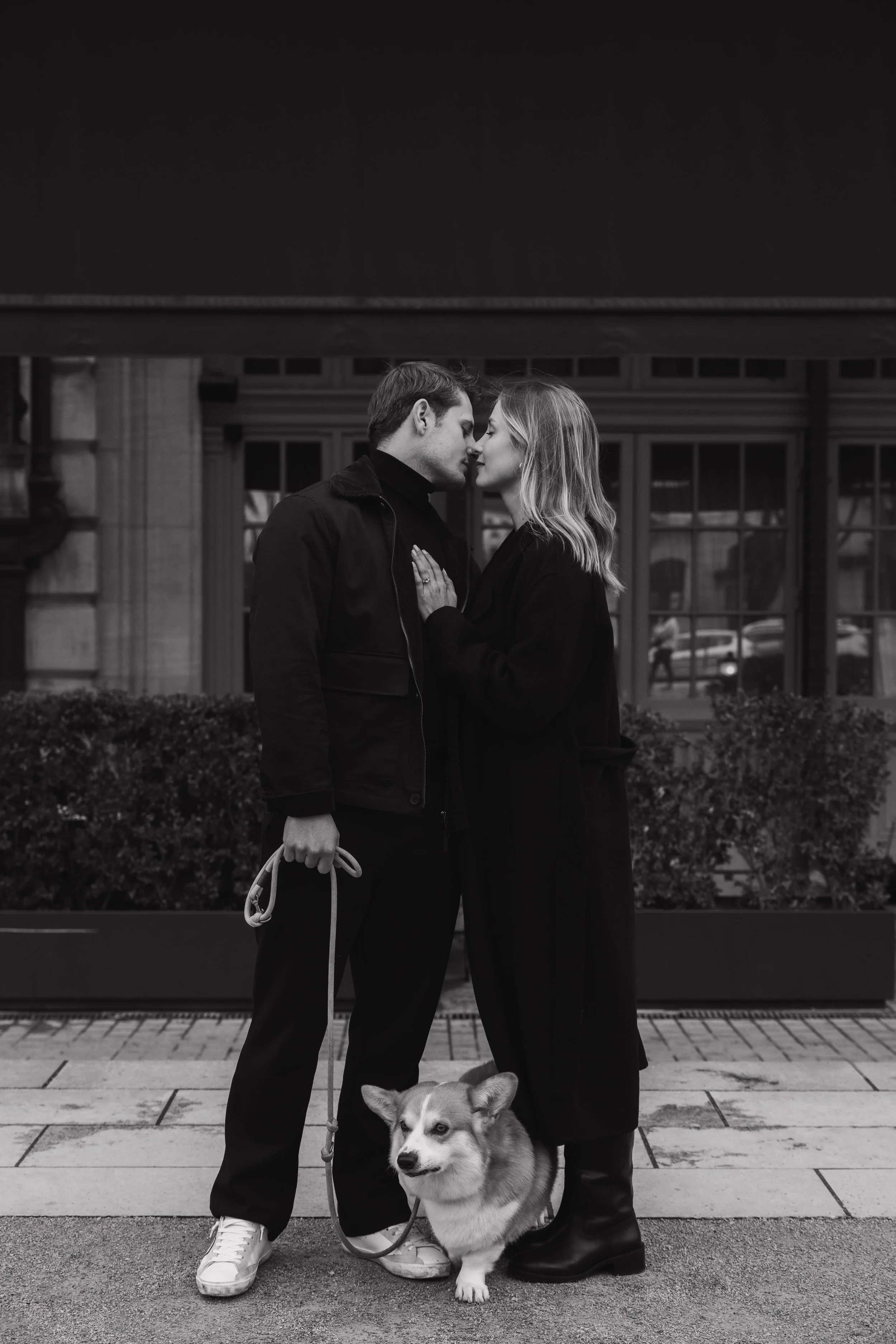 Un couple s'apprête à s'embrasser, elle pose sa main sur sa poitrine et il tient une laisse avec un chien de race Pembroke Welsh Corgi à ses pieds, en noir et blanc, en ville. session engagement couple Lyon 6eme, brotteaux, photographe mariage