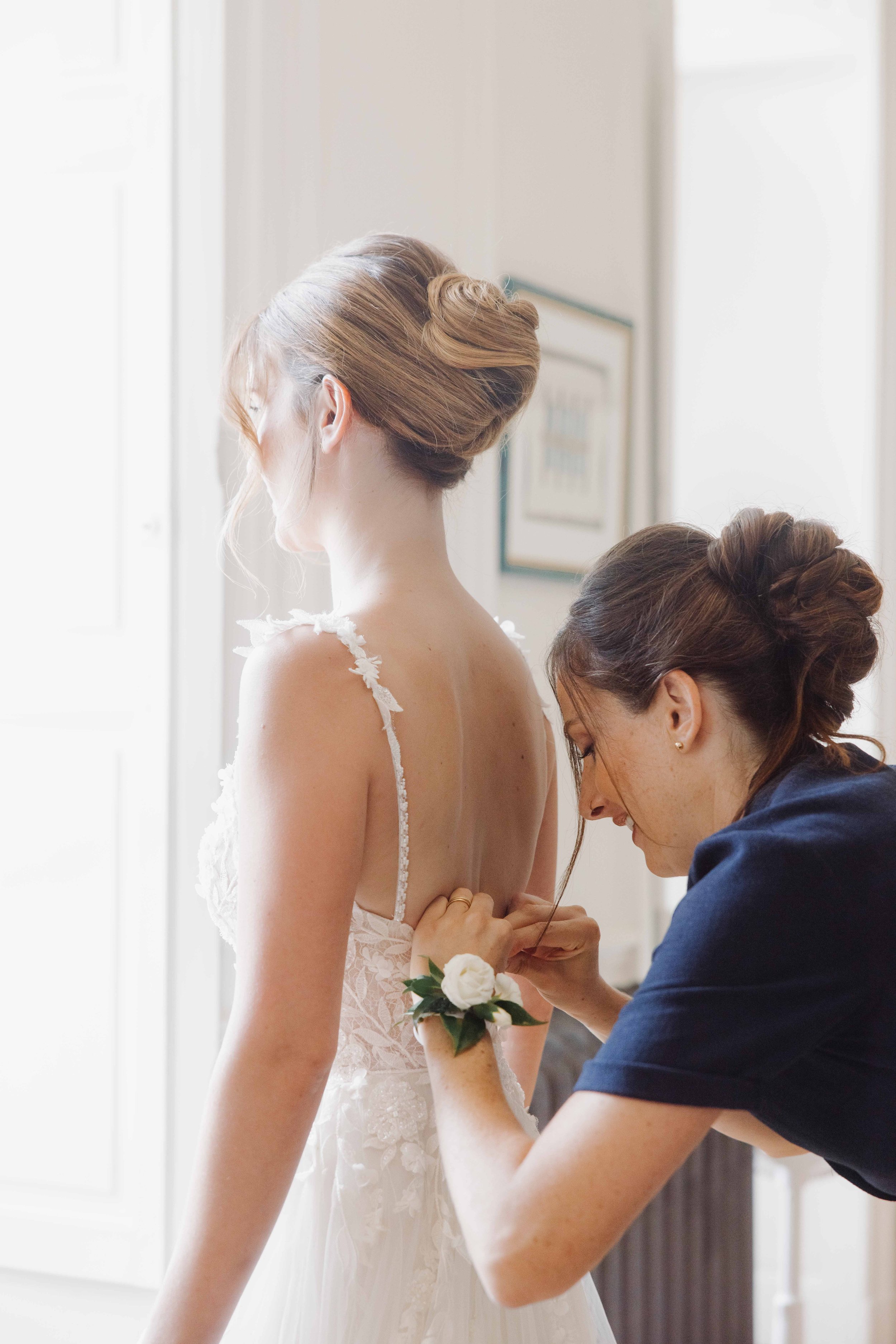 Une femme ajuste la robe de mariée d'une autre femme, dans une pièce lumineuse. Mariage chateau de champ renard, Blacé, beaujolais, Lyon, destination wedding. préparatifs 