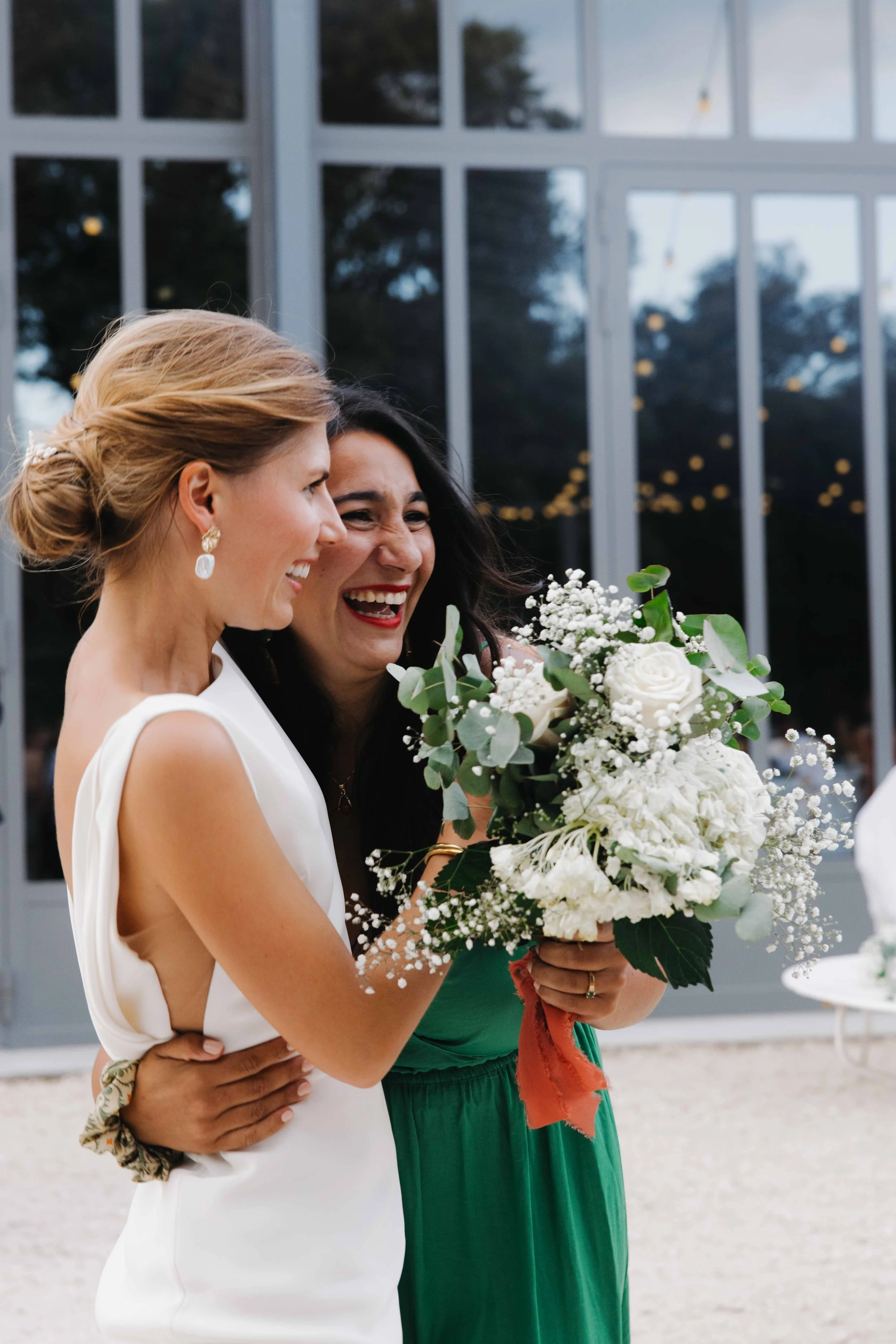 Deux femmes sourient et se câlinent lors d'une cérémonie, l'une tenant un bouquet de fleurs blanches, tout en étant proches d'une grande fenêtre avec des lumières dans le fond. Domaine du bijoutier à grignan, drome provençal, coucher de soleil