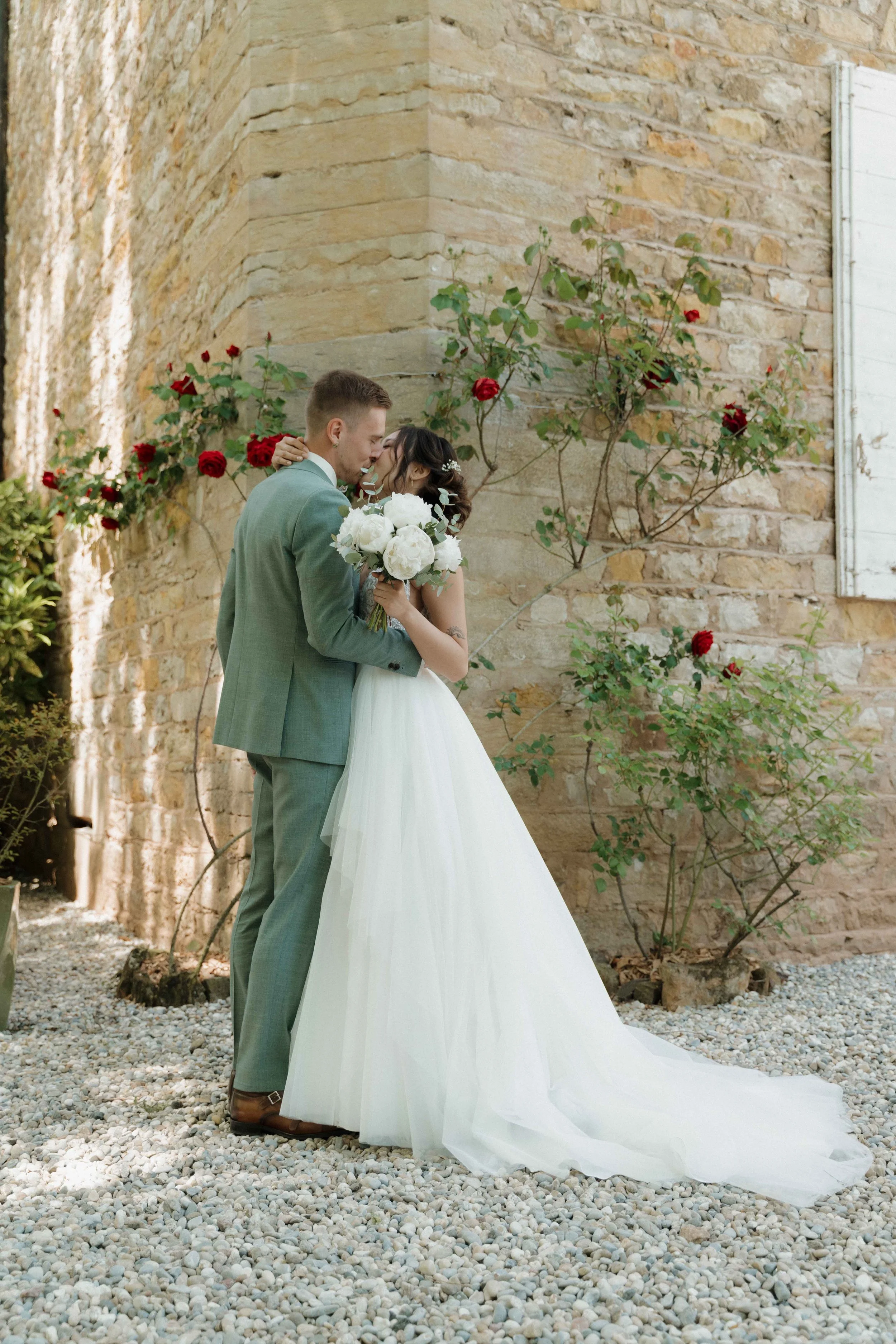 Un couple de mariés en train de s'embrasser devant un mur en pierre avec des roses rouges grimpantes. Domaine de tourieux à Savigny dans le beaujolais à Lyon. Mariage cérémonie laïque, photographe Lyon haut de gamme, duo photographe, argentique.