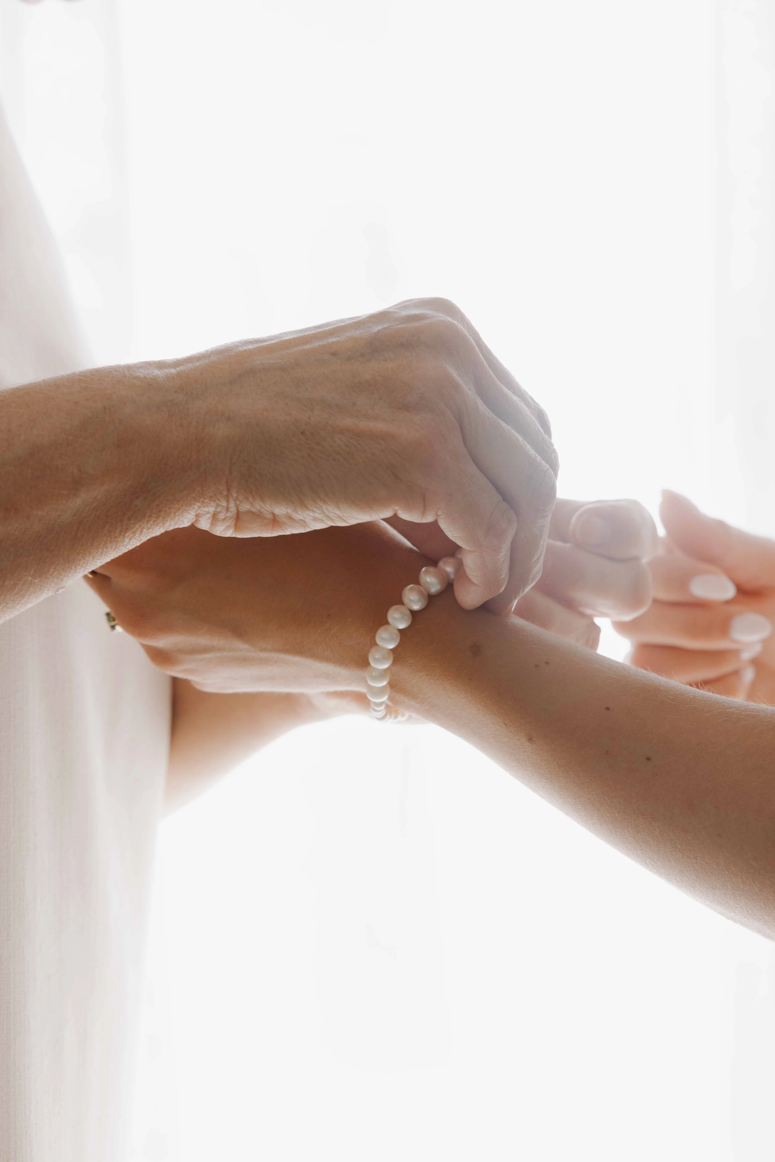 Gros plan sur deux mains tenant un bracelet de perles, une personne aidant une autre à le mettre, lumière naturelle en arrière-plan. Photographe mariage Isère, ferme du rocher, cérémonie civil et laïque. Bordeaux et Lyon et destination wedding. 