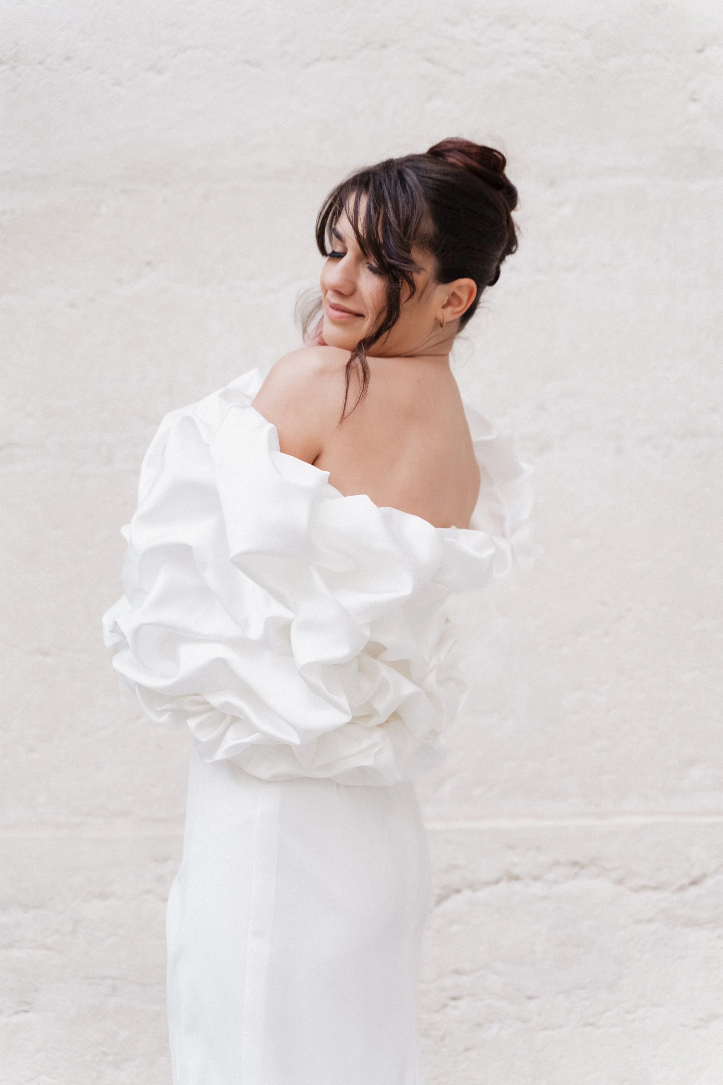 Femme en robe blanche avec une peau nue au dos, souriante, portant une robe à volants, devant un mur beige  à l'hôtel dieu à Lyon 2ème, à l'intercontinental, session engament, photographe mariage duo haut de gamme.