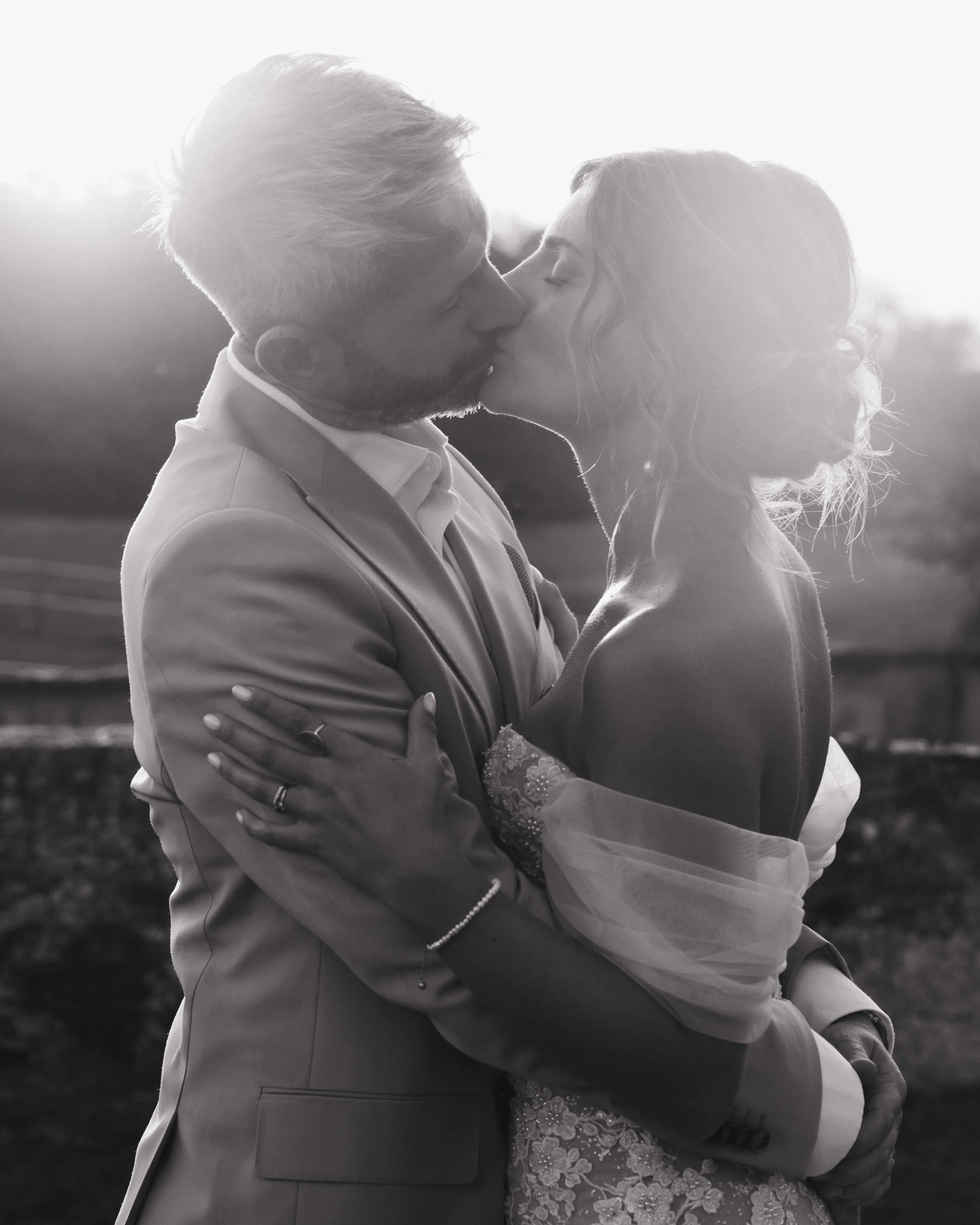 Un couple en costume de mariage s'embrasse lors d'une séance photographique en extérieur, en noir et blanc. Mariage à la ferme du rocher en Isère, robe de mariées, coucher de soleil, photos de couples. Photographe en duo haut de gamme et destination 
