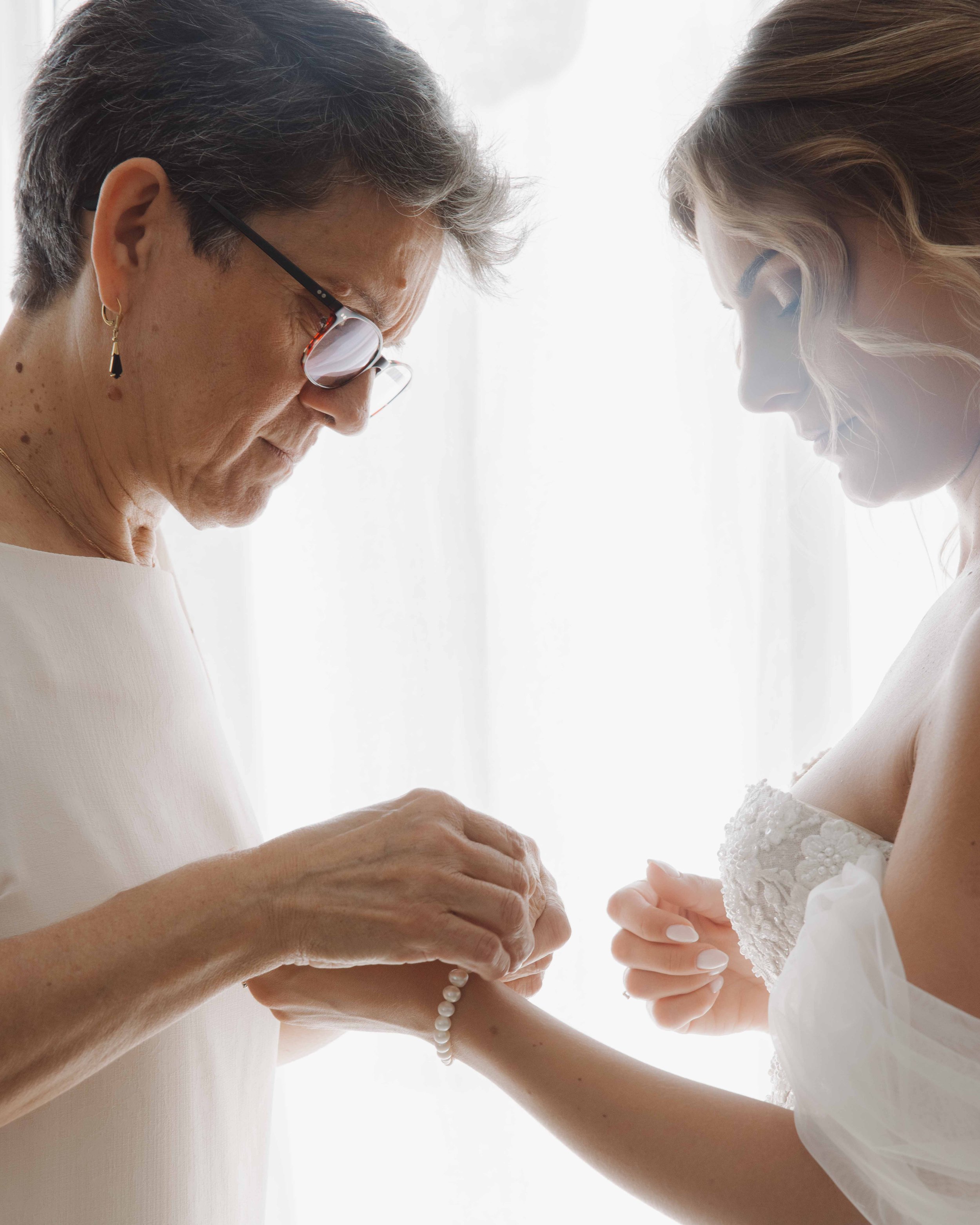Une femme plus âgée aide une jeune femme à mettre un bracelet de perles, probablement lors d'une préparation pour un mariage.