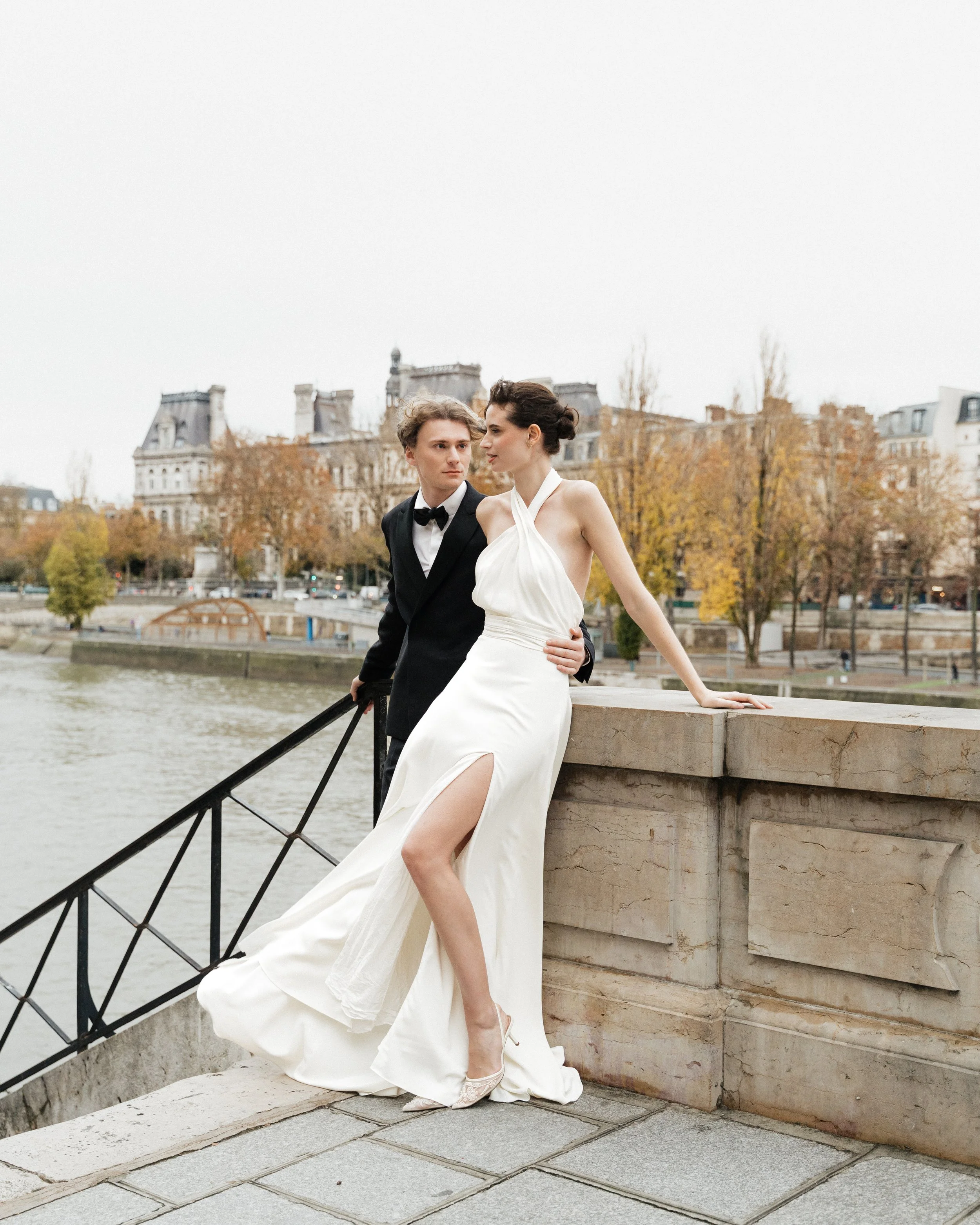 Un couple en vêtements de mariage posé sur un quai au bord de la rivière à Paris, avec des arbres aux feuilles automnales et des bâtiments en arrière-plan. Session engagement paris, Louvre, mariage haut de gamme, destination wedding. Robe de mariées 