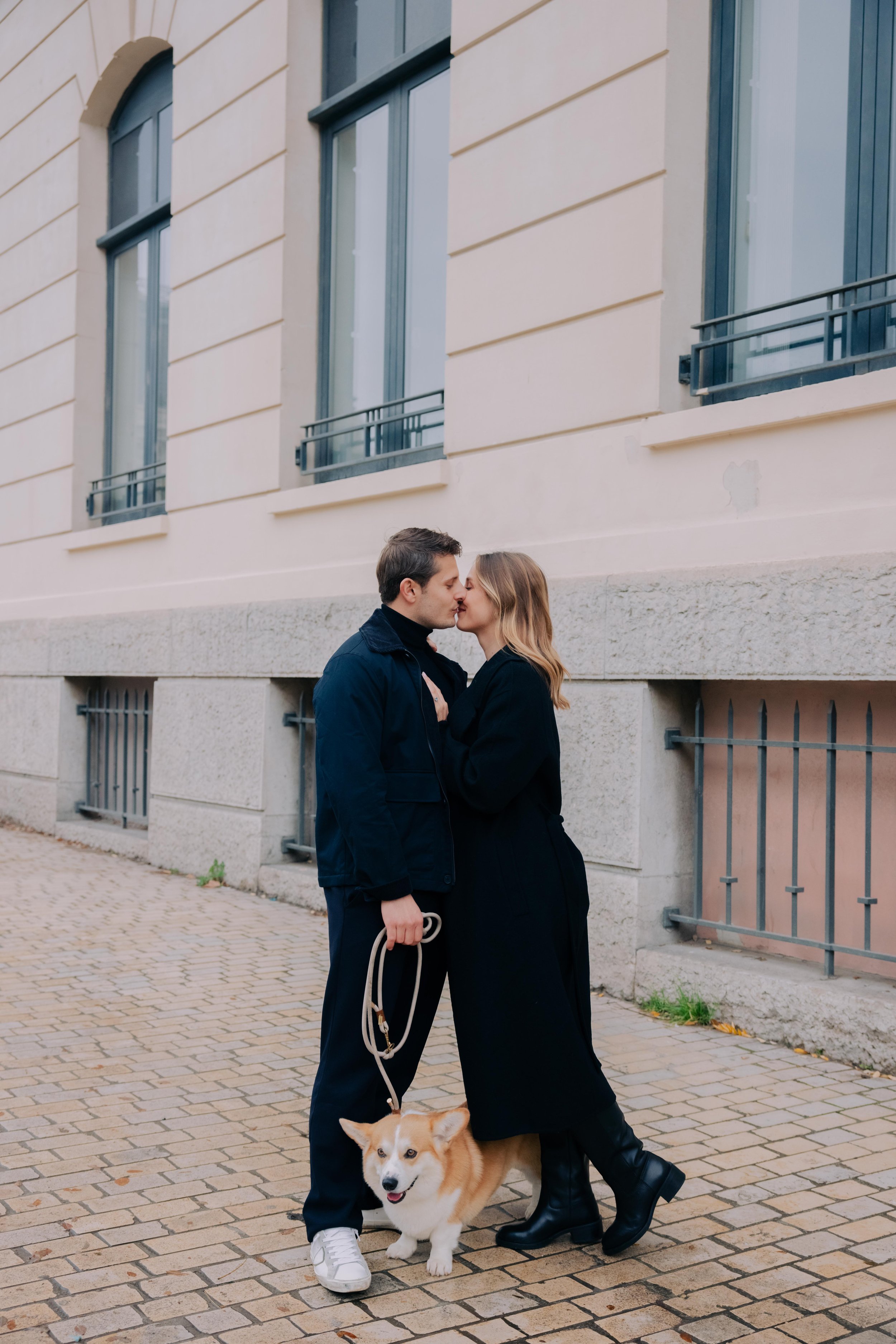 Un couple s'embrassant devant un bâtiment beige avec fenêtres. Le couple tient une chienne corgi beige avec blanc. La femme porte une robe noire et des bottes noires, l'homme porte une veste foncée et des baskets blanches. session engagement couple 