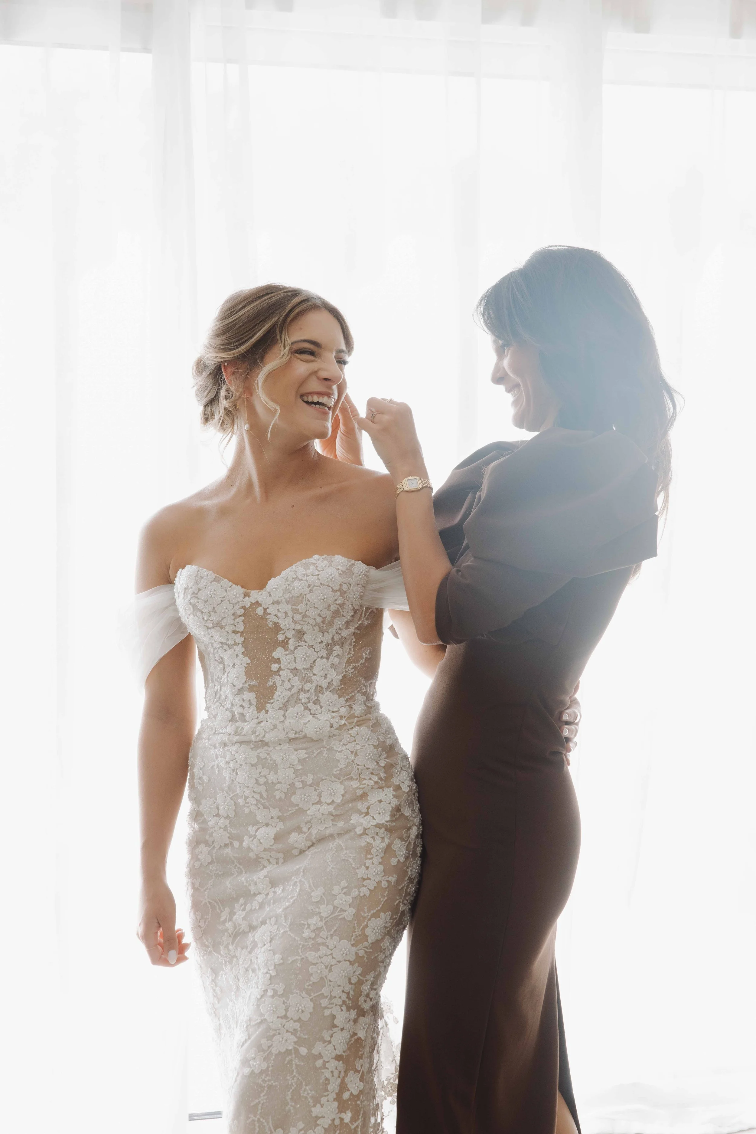 Deux femmes, une en robe de mariée et l'autre en robe noire, sourient et se maquillent à l'arrière d'une fenêtre en lumière naturelle. Photographe mariage Isère, ferme du rocher, cérémonie civil et laïque. Bordeaux et Lyon et destination wedding. 