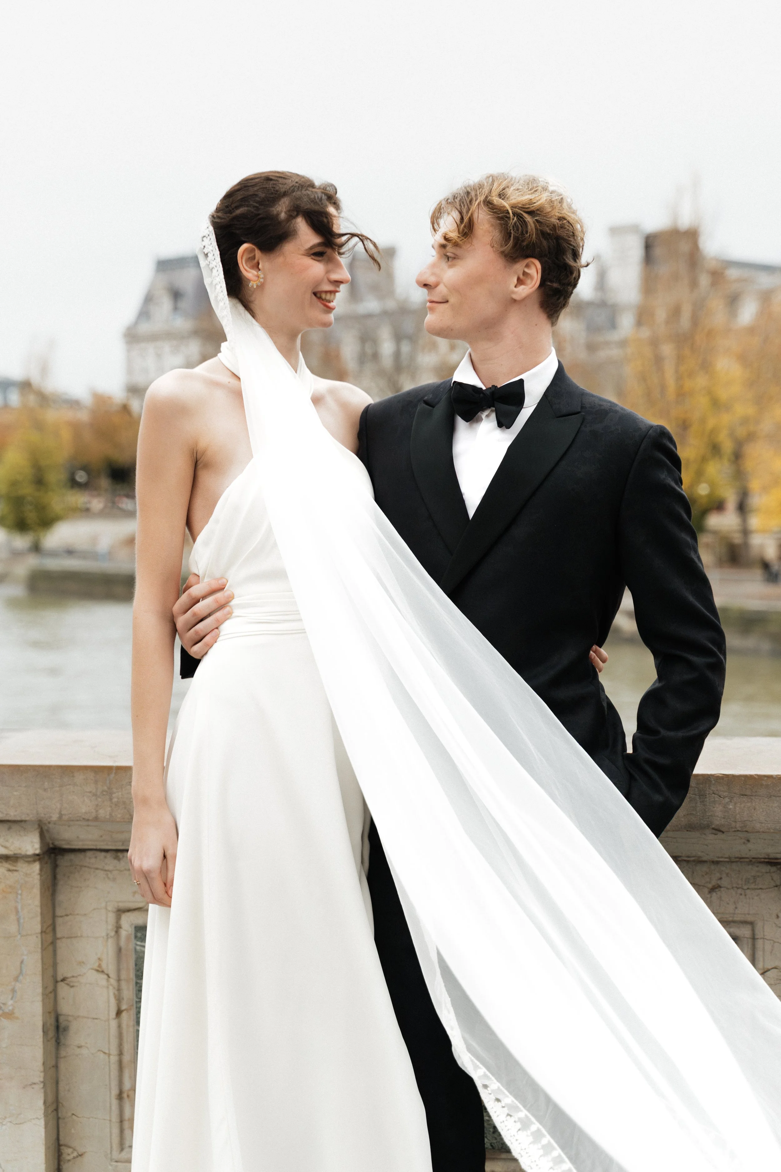 Un couple en vêtements de mariée  , se regarde tendrement devant un reflet d'eau avec des bâtiments en arrière-plan, en automne. Session engagement paris, Louvre, mariage haut de gamme, destination wedding. Robe de mariées, madame a dit oui