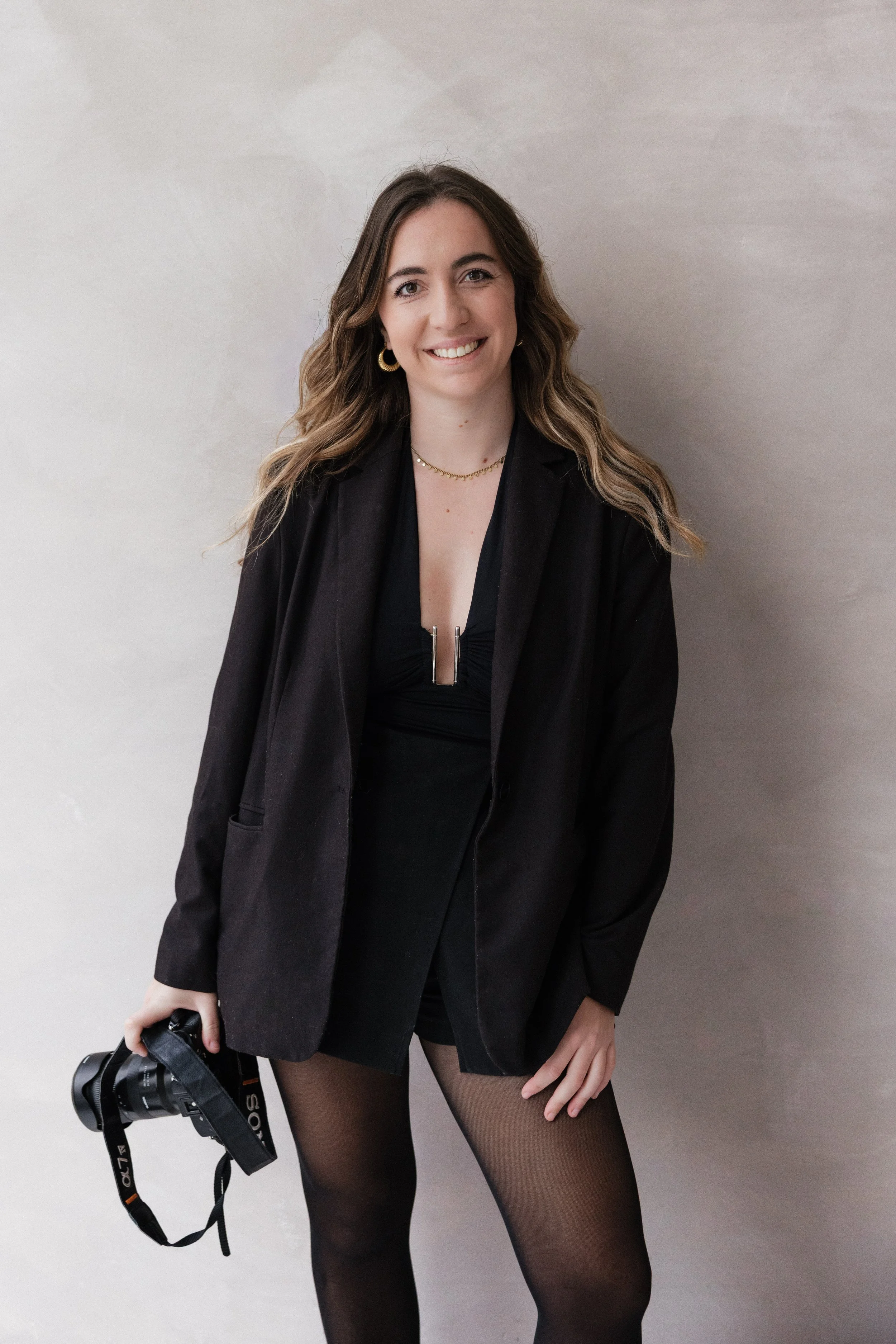 Une femme souriante tenant un appareil photo, portant une veste noire, un haut noir, des collants noirs, des boucles d'oreilles dorées et un collier, posant contre un mur clair. Photographe Lyon, bordeaux, France, Europe, film & digital