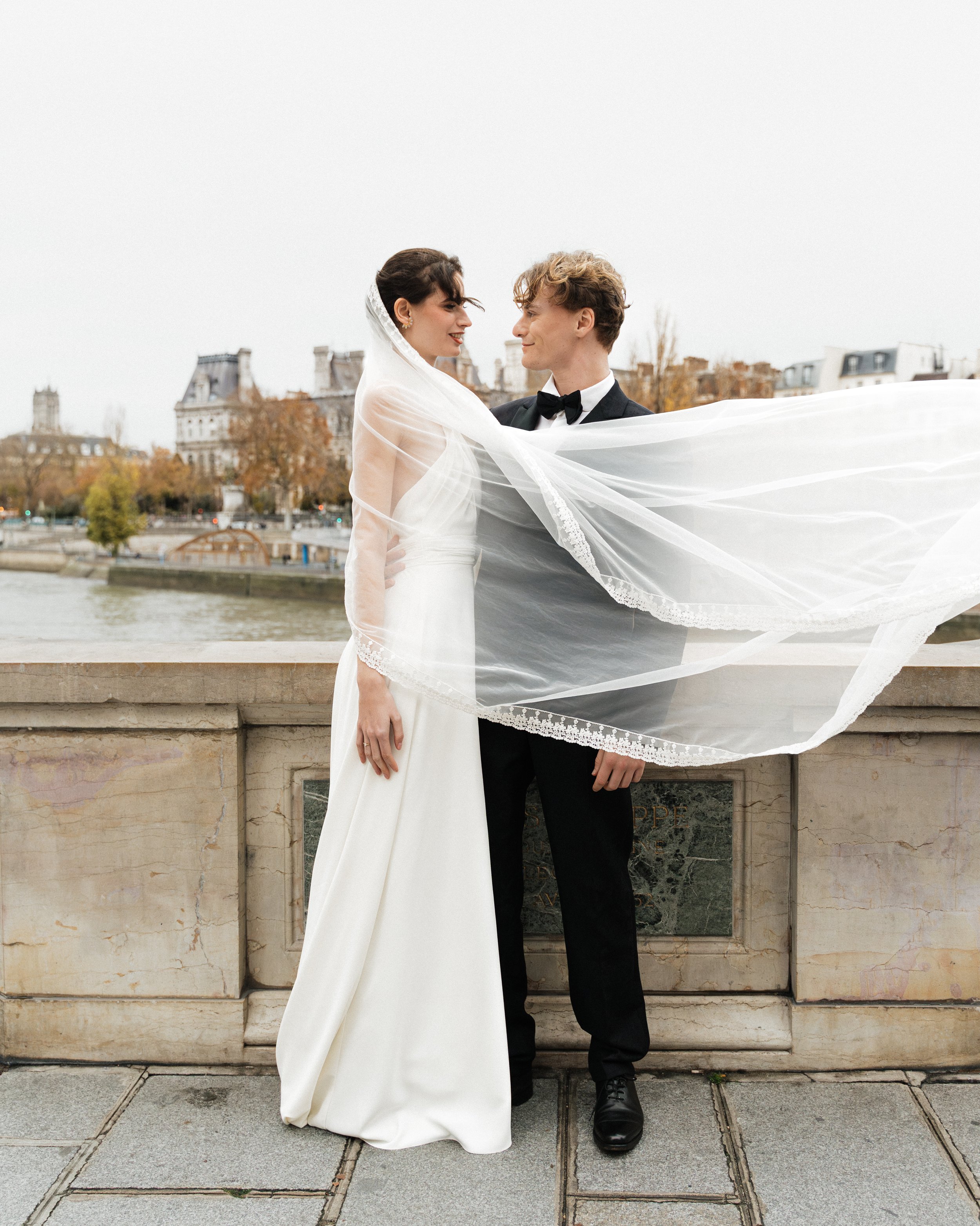 Un couple marié en vêtements de mariage se regarde tendrement devant la Seine à Paris, avec des bâtiments historiques en arrière-plan, en automne. Session engagement paris, Louvre, mariage haut de gamme, destination wedding. Robe de mariées 