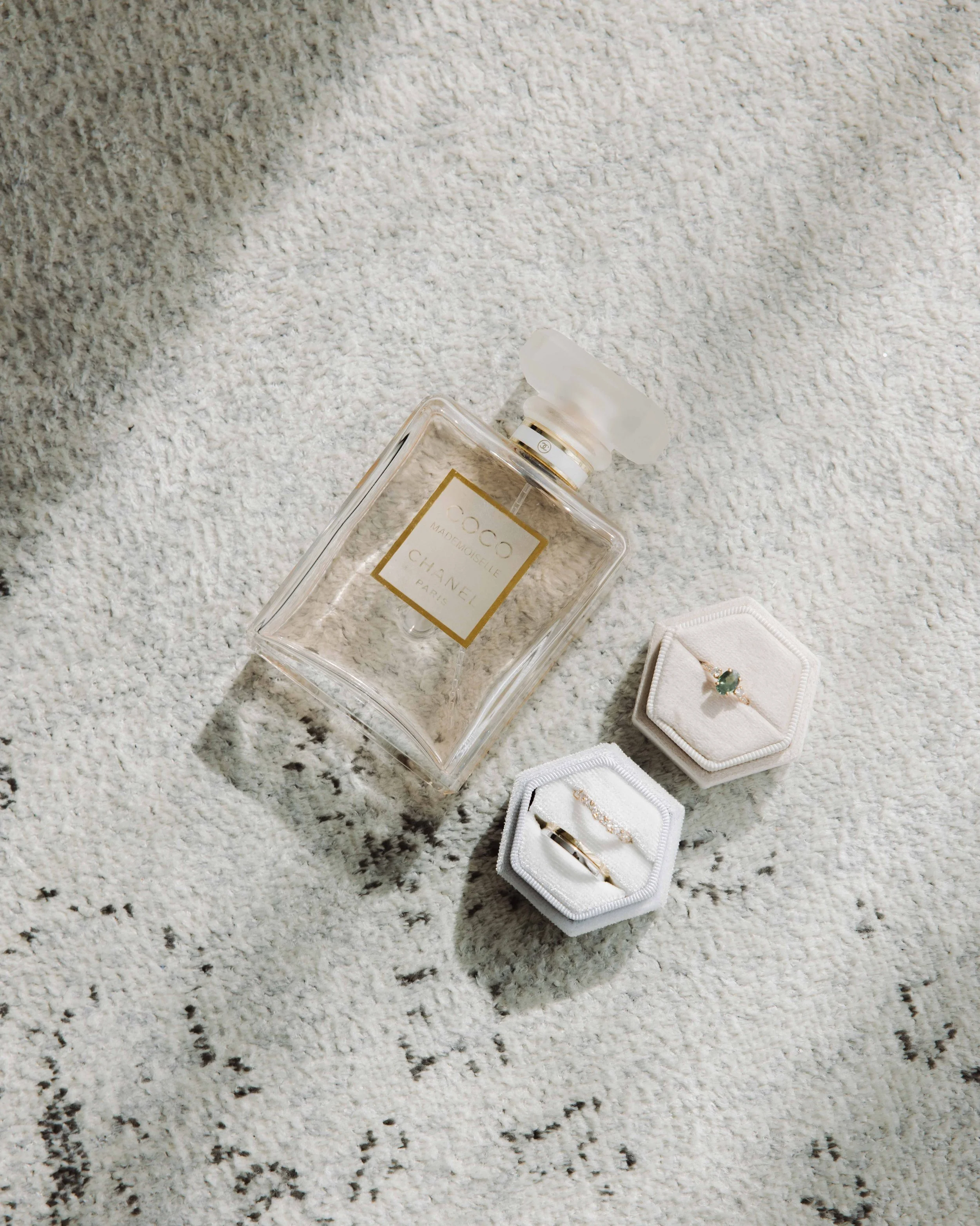Une bouteille de parfum Chanel Coco Mademoiselle et deux bagues dans des boîtes hexagonales, posés sur un tapis blanc. Photographe mariage Isère, ferme du rocher, cérémonie civil et laïque. Bordeaux et Lyon et destination wedding. Photos de couple 