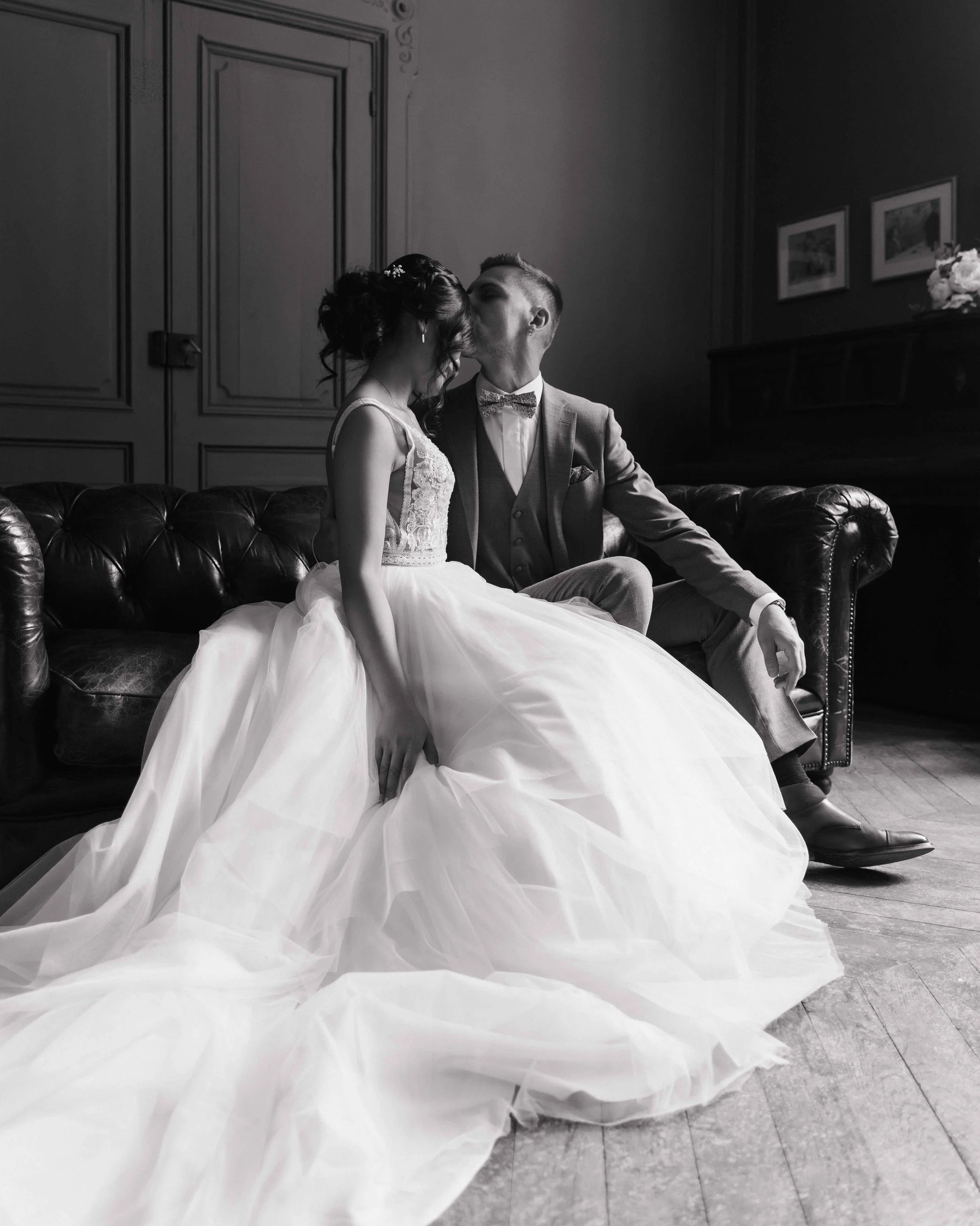 Un couple en mariage s'embrassant sur un canapé en cuir dans une pièce élégante, la mariée portant une robe blanche volumineuse, l'homme vêtu d'un costume gris et d'une cravate à motifs. Domaine de tourieux à Savigny dans le beaujolais à Lyon 