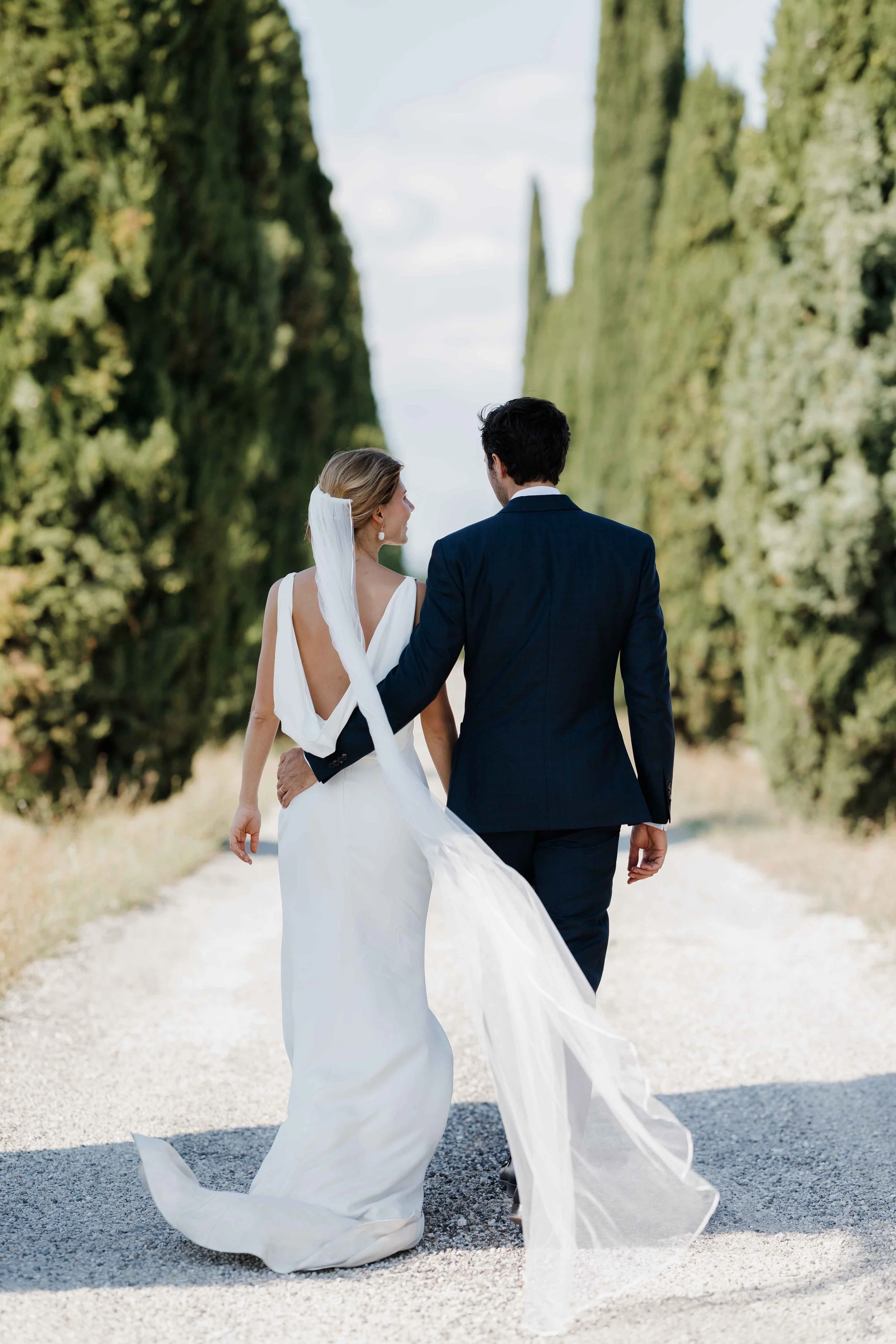 Un couple en mariage marche sur une route bordée de grands arbres verts. Domaine du bijoutier à grignan, drome provençal, coucher de soleil, session couple, mariage religieux, cérémonie religieuse 