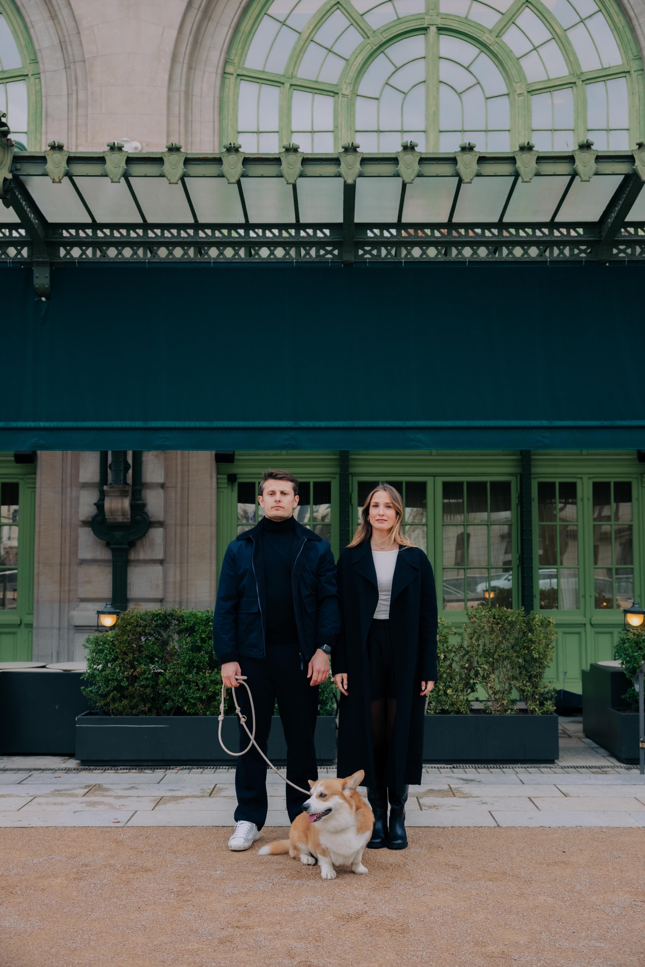 Un homme et une femme posant devant un bâtiment avec des fenêtres vertes, accompagnés d'un chien de race Pembroke Welsh Corgi. session engagement couple Lyon 6eme, brotteaux, photographe mariage bordeaux et Lyon et destination wedding