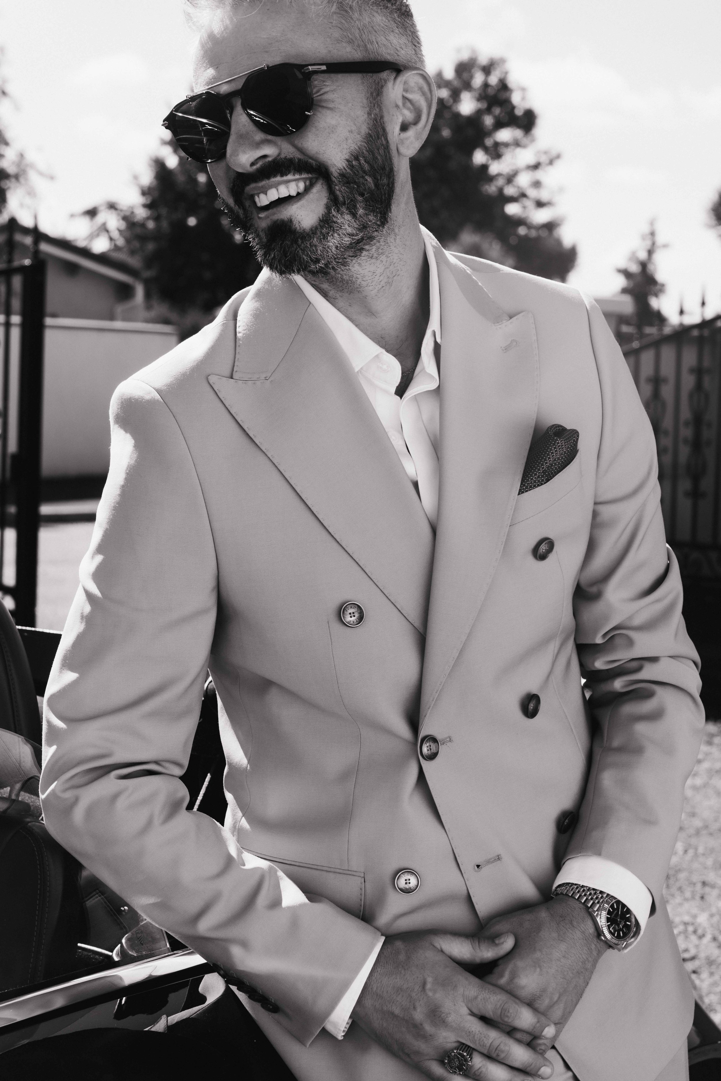 Homme souriant portant un costume double-breast, lunettes de soleil et montre. Photographe mariage Isère, ferme du rocher, cérémonie civil et laïque. Bordeaux et Lyon et destination wedding. Photos de couple, session couples. Préparatifs 