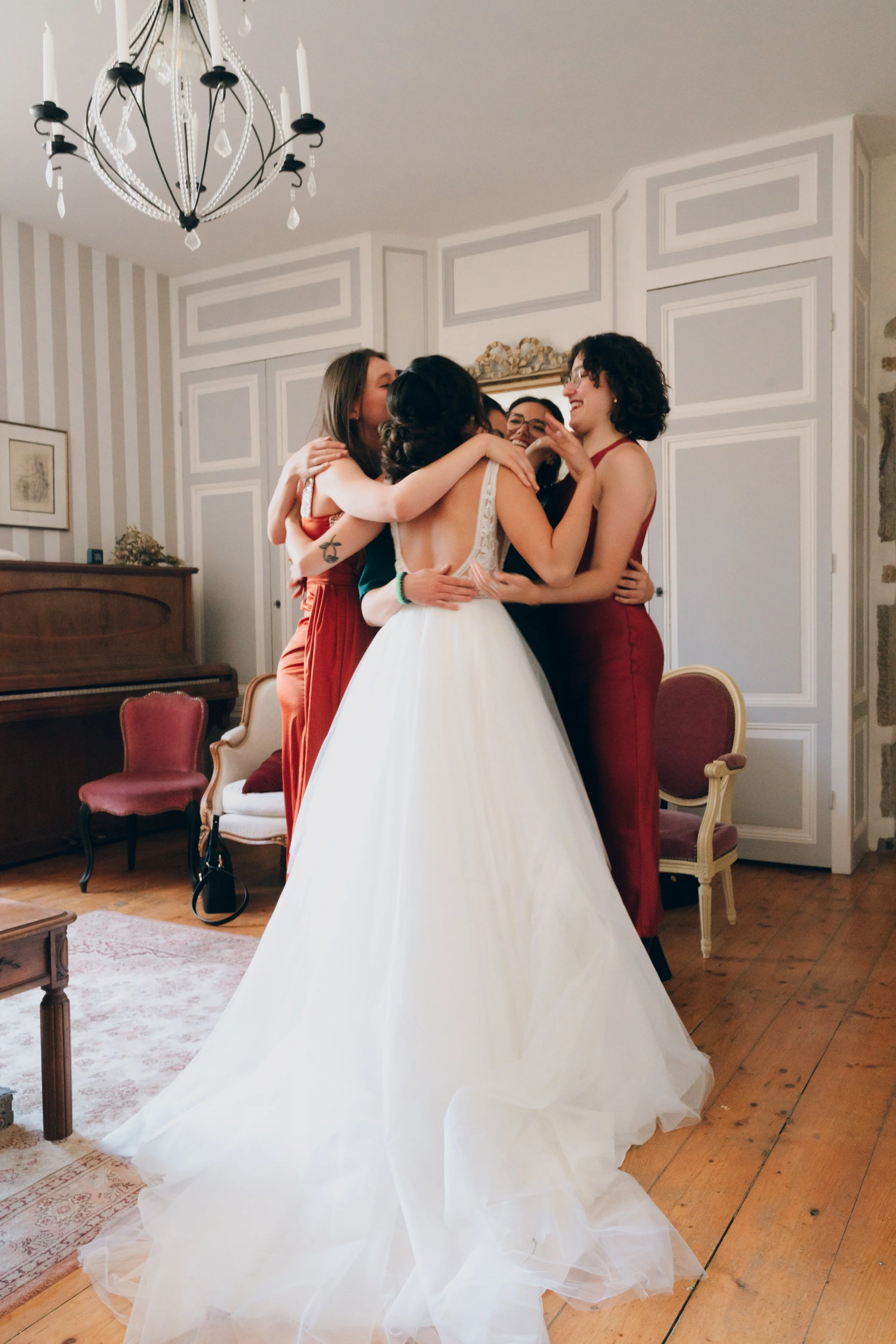 Une mariée en robe blanche entourée de quatre femmes en robes rouges dans une pièce décorée élégamment, partageant une étreinte joyeuse. Domaine de tourieux à Savigny dans le beaujolais à Lyon. Mariage cérémonie laïque, photographe Lyon haut de gamme