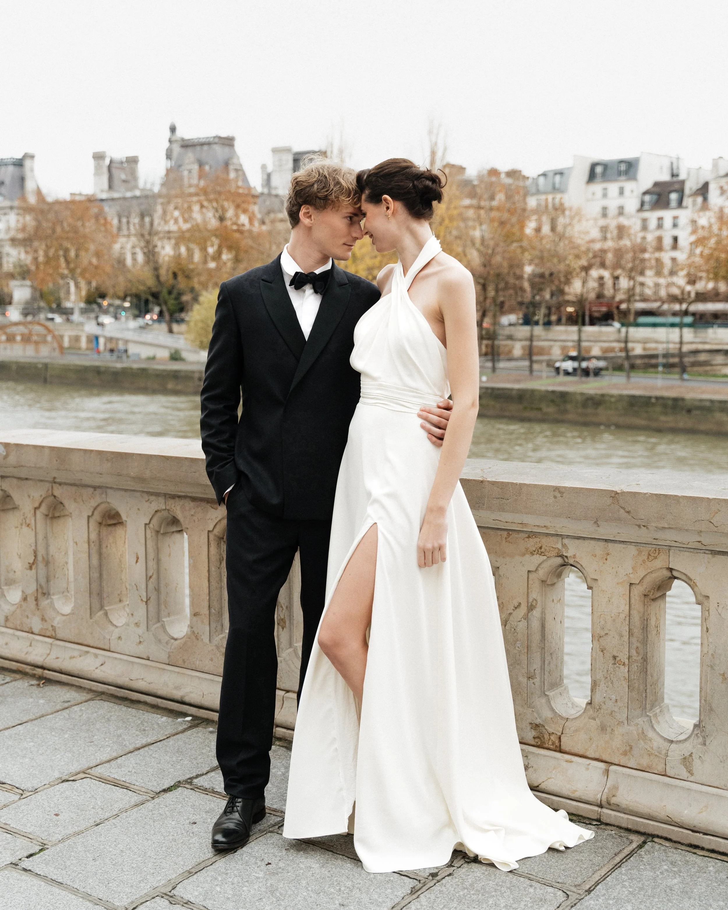 Un couple en vêtements formels, un homme en smoking et une femme en robe de mariage, se tenant sur une terrasse au bord d'une rivière, avec des bâtiments parisiens en arrière-plan. Session engagement paris, Louvre, madame a dit oui 