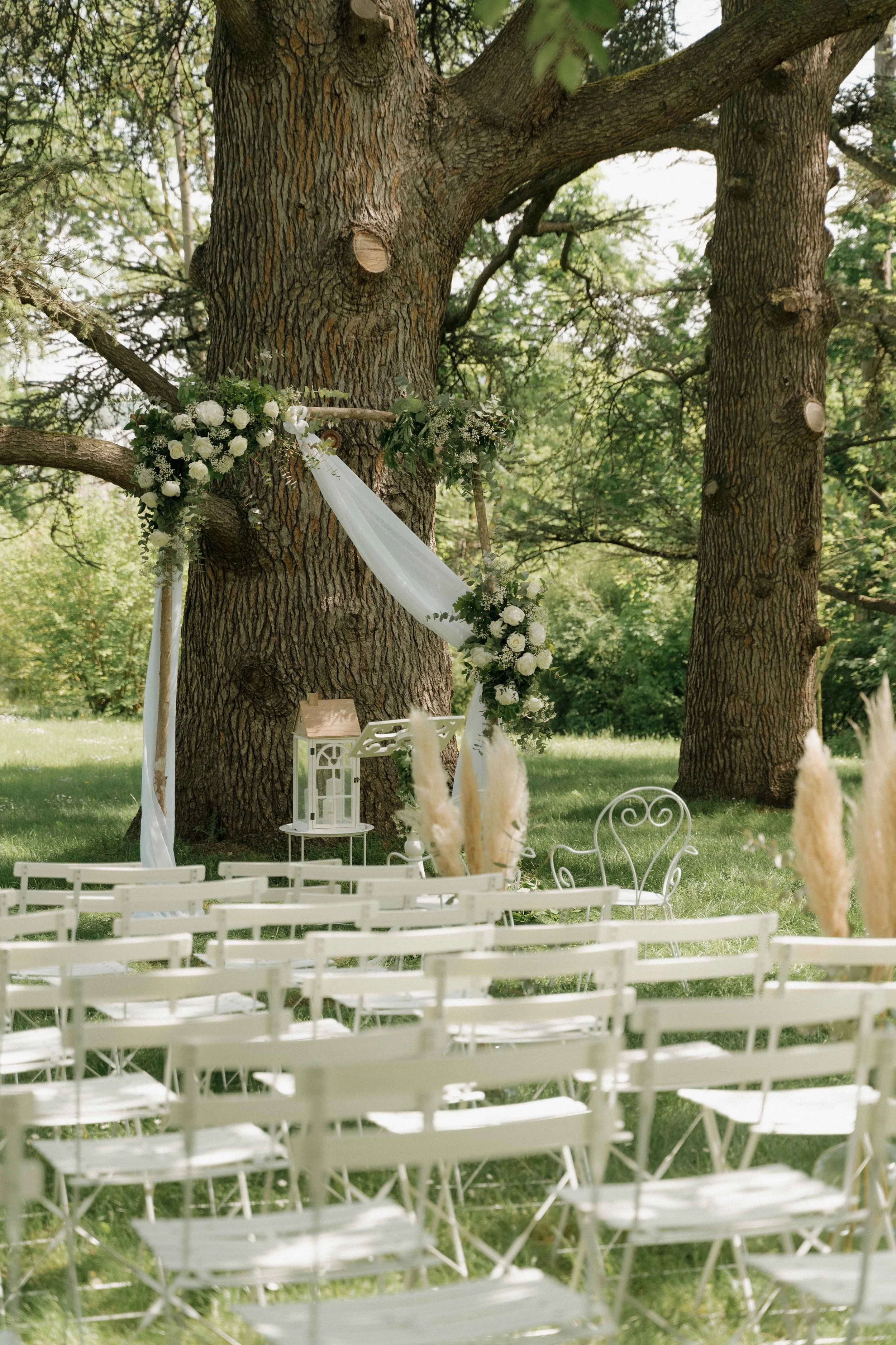 Un lieu de mariage en plein air avec une arche décorée de fleurs blanches et de tissu blanc, sous de grands arbres, avec des chaises blanches alignées pour les invités. Domaine de tourieux à Savigny dans le beaujolais à Lyon. Mariage cérémonie laïque
