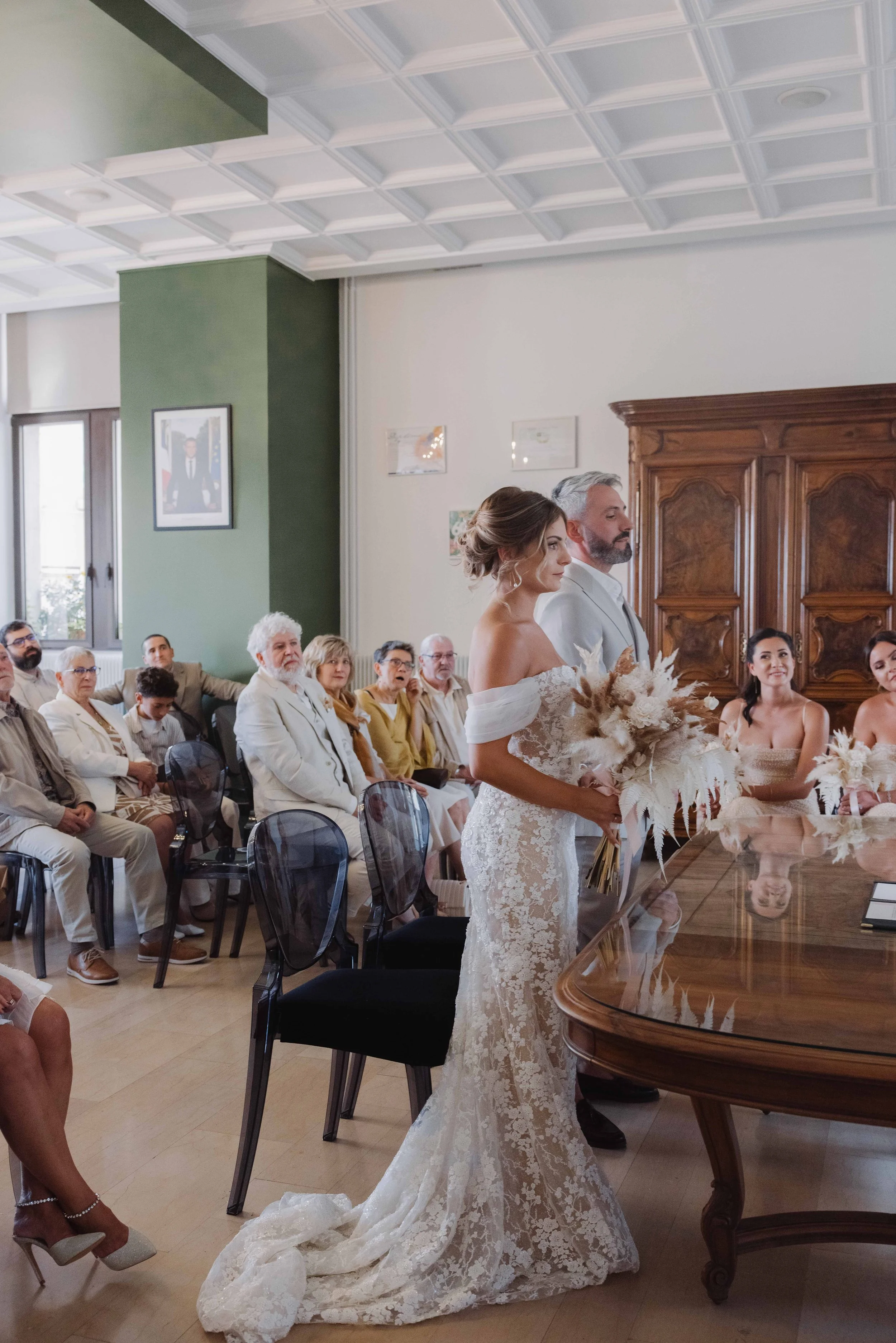 Mariée en robe de mariage ajustée en dentelle, tenant un bouquet de fleurs naturelles, lors de la cérémonie, entourée de ses invités dans une salle de mariage élégante. Photographe mariage Isère, ferme du rocher, cérémonie civil et laïque. Lyon 