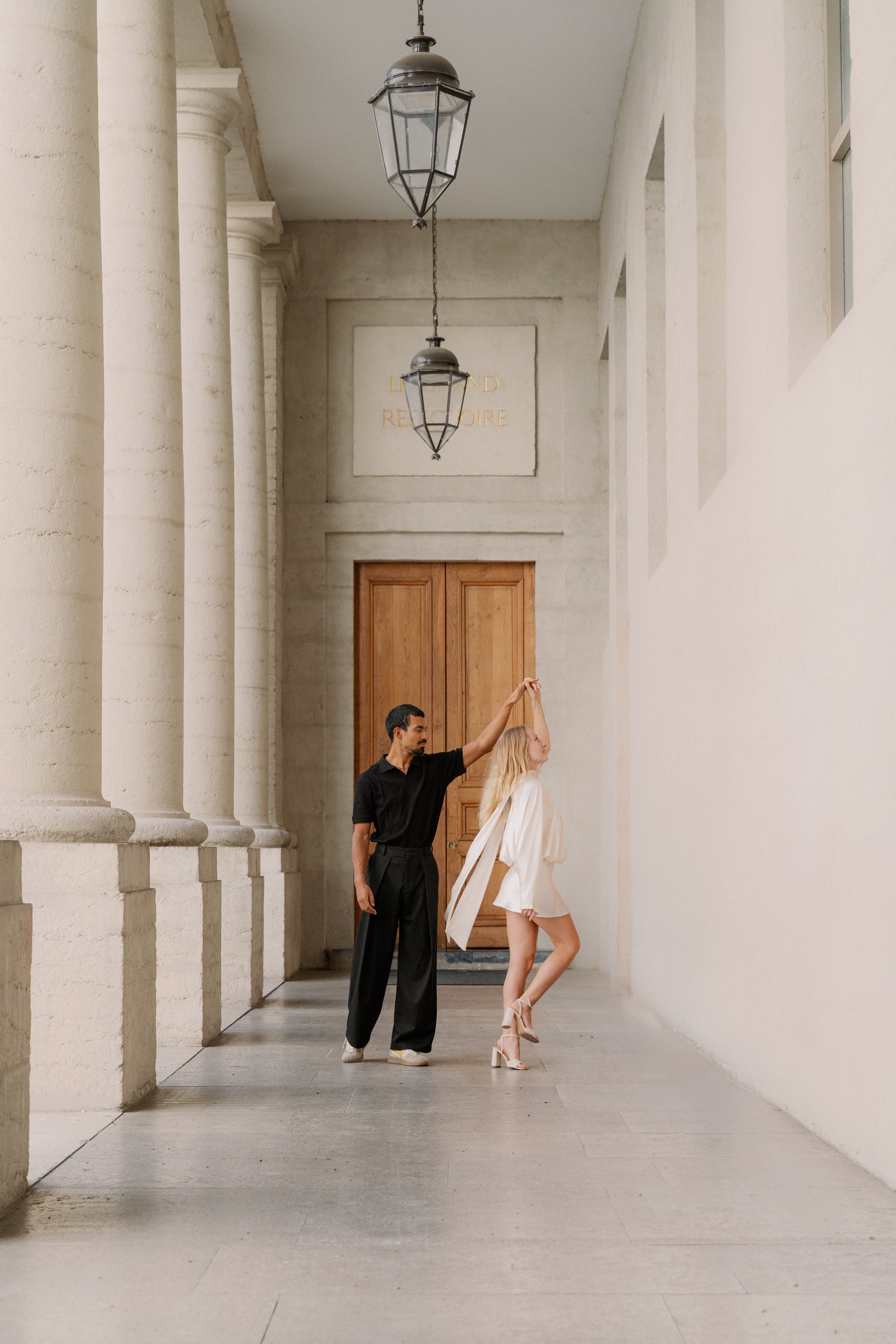 Un homme noir danse avec une femme blonde en robe blanche dans un couloir avec colonnes et lampes suspendues à l'hôtel dieu à Lyon 2ème, à l'intercontinental, session engament, photographe mariage duo haut de gamme.