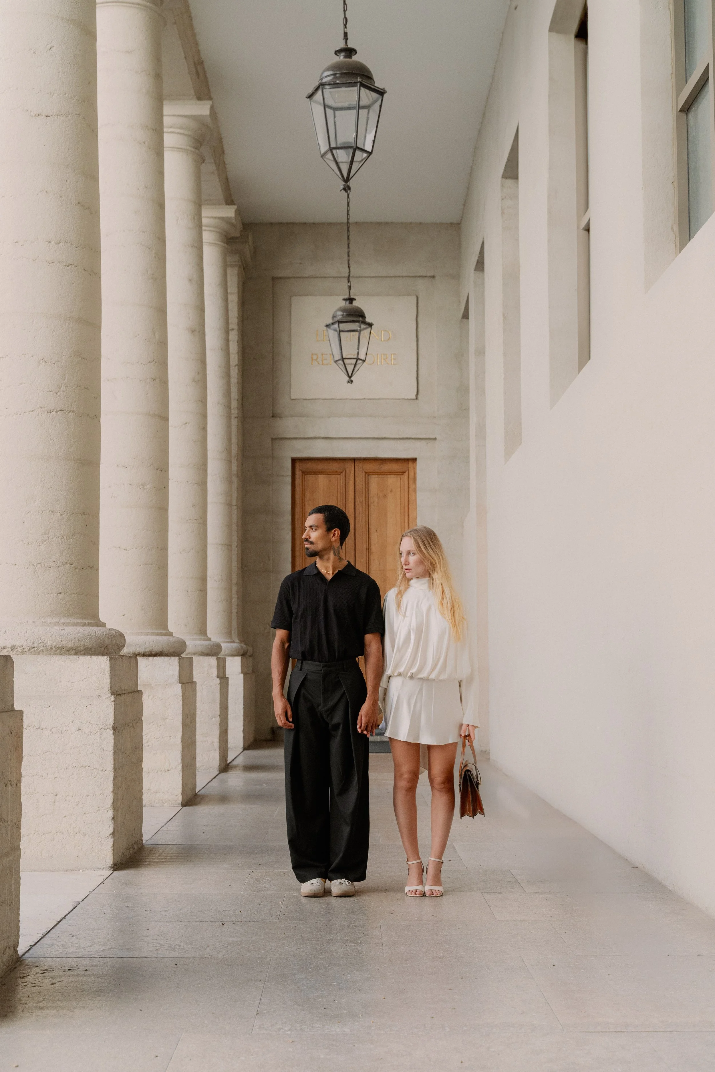 Un couple marche main dans la main dans un couloir lumineux avec des colonnes et des lampes suspendues à l'hôtel dieu à Lyon 2ème, à l'intercontinental, session engament, photographe mariage duo haut de gamme.