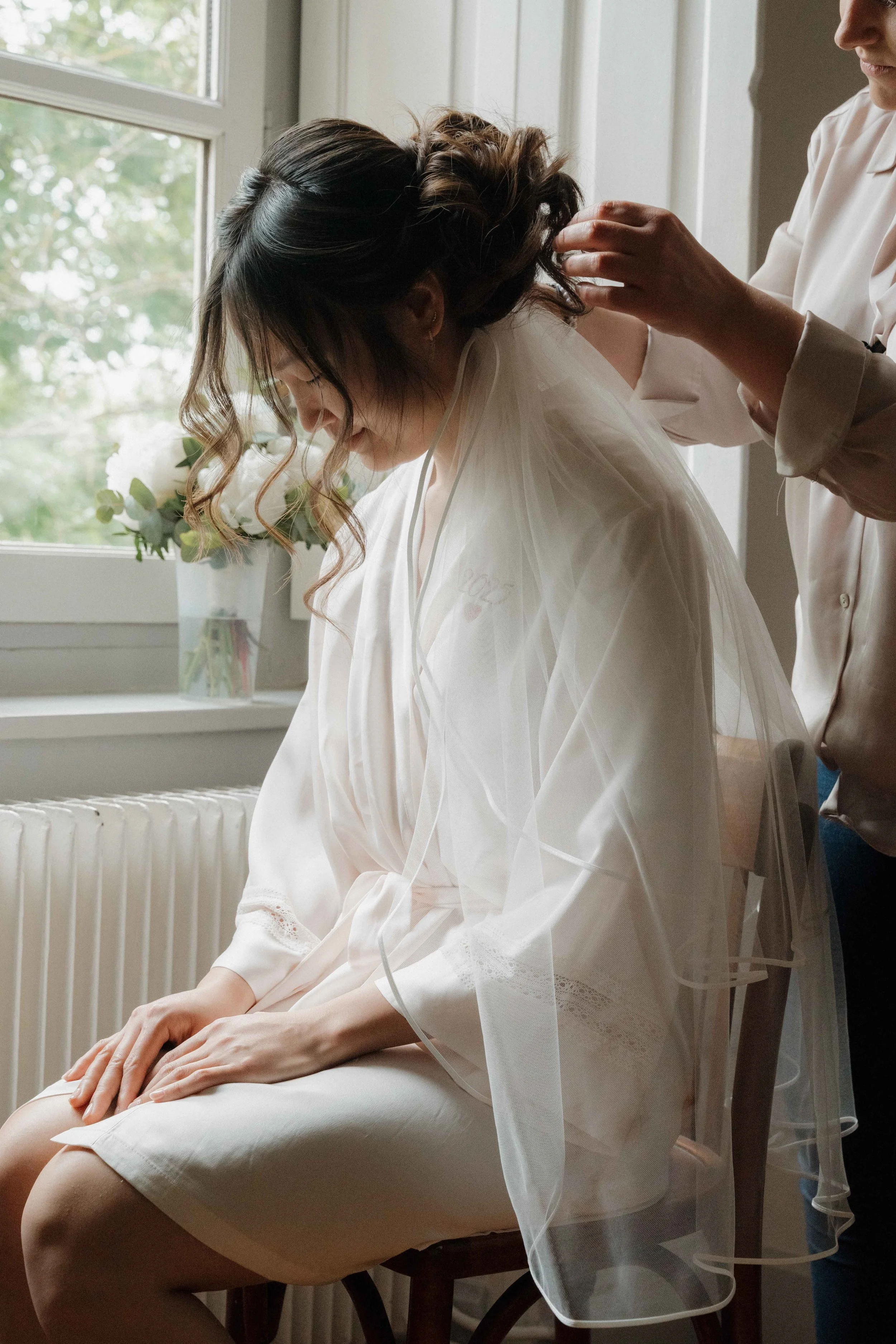 Une femme en robe de satin blanc, assise sur une chaise, en train de se faire coiffer par une autre personne, dans une pièce près d'une fenêtre avec une plante en vase sur le rebord. Domaine de tourieux à Savigny dans le beaujolais à Lyon. Mariage