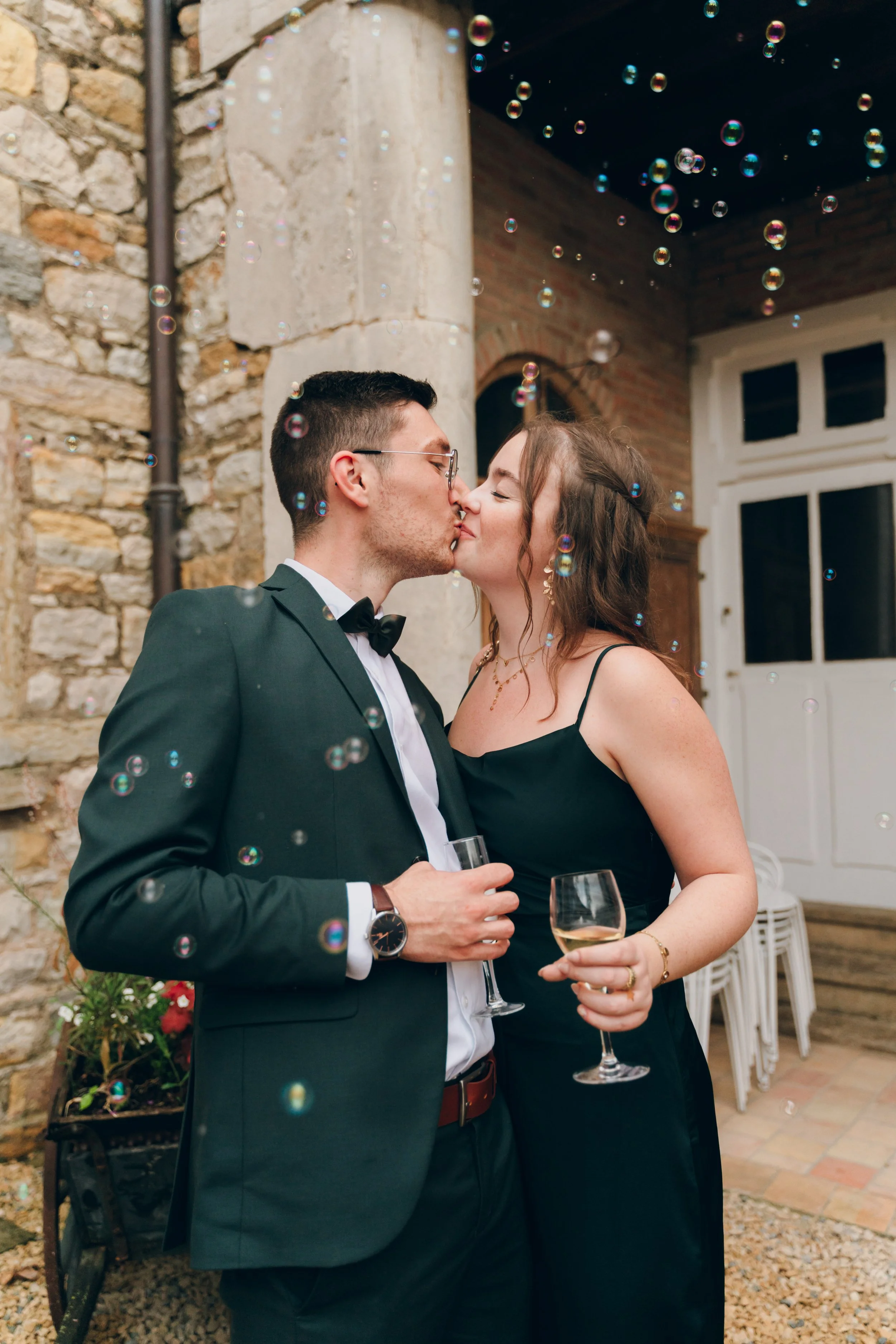 Un couple en tenue formelle s'embrasse en se tenant par la taille lors d'une fête ou d'un mariage, avec des bulles flottantes dans l'air et une ambiance chaleureuse en arrière-plan. Domaine de tourieux à Savigny dans le beaujolais à Lyon. 