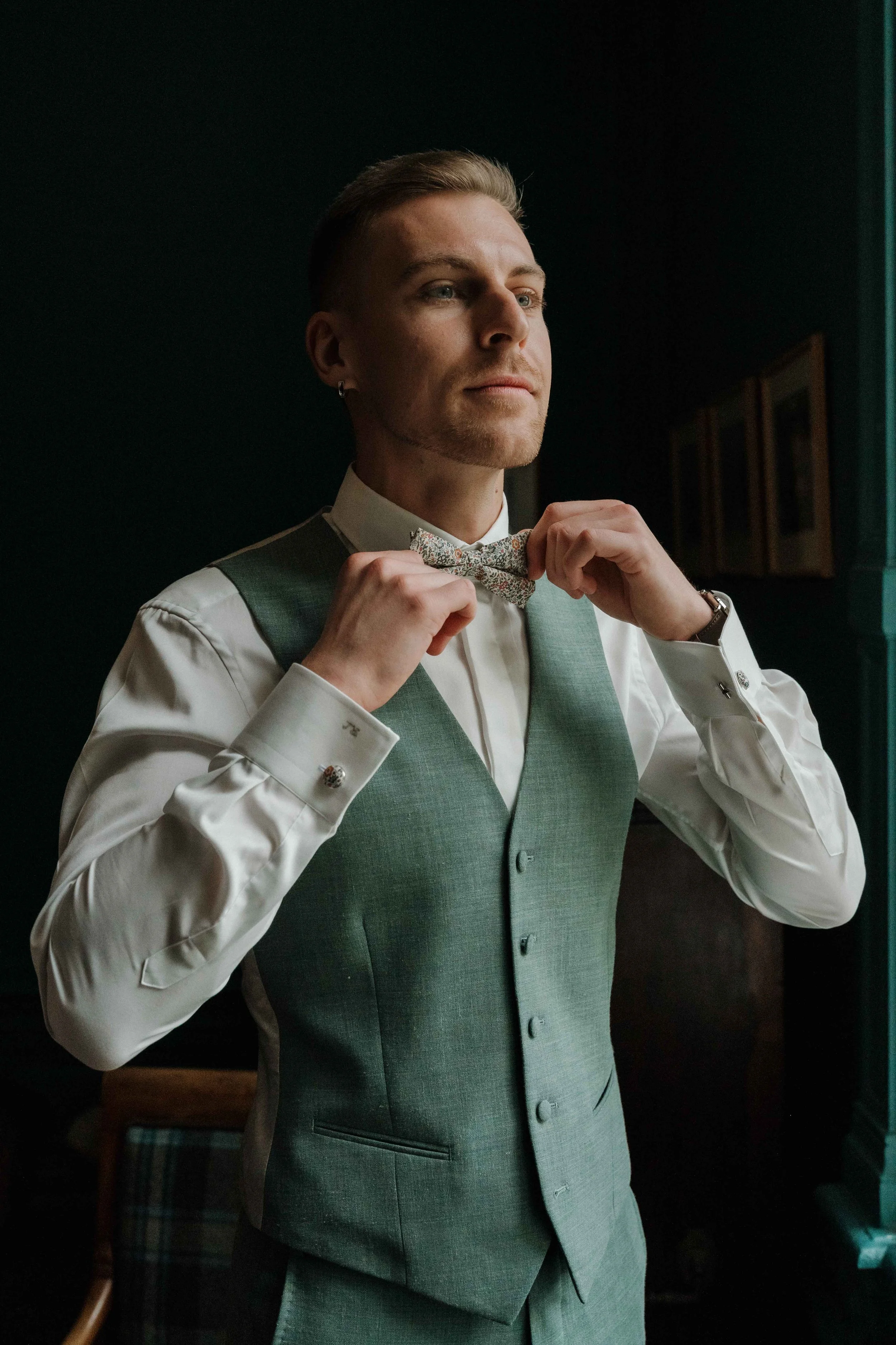 Un homme élégant ajuste un nœud papillon à l'intérieur, portant une veste vert olive et une chemise blanche. Domaine de tourieux à Savigny dans le beaujolais à Lyon. Mariage cérémonie laïque, photographe Lyon haut de gamme, duo photographe, film 