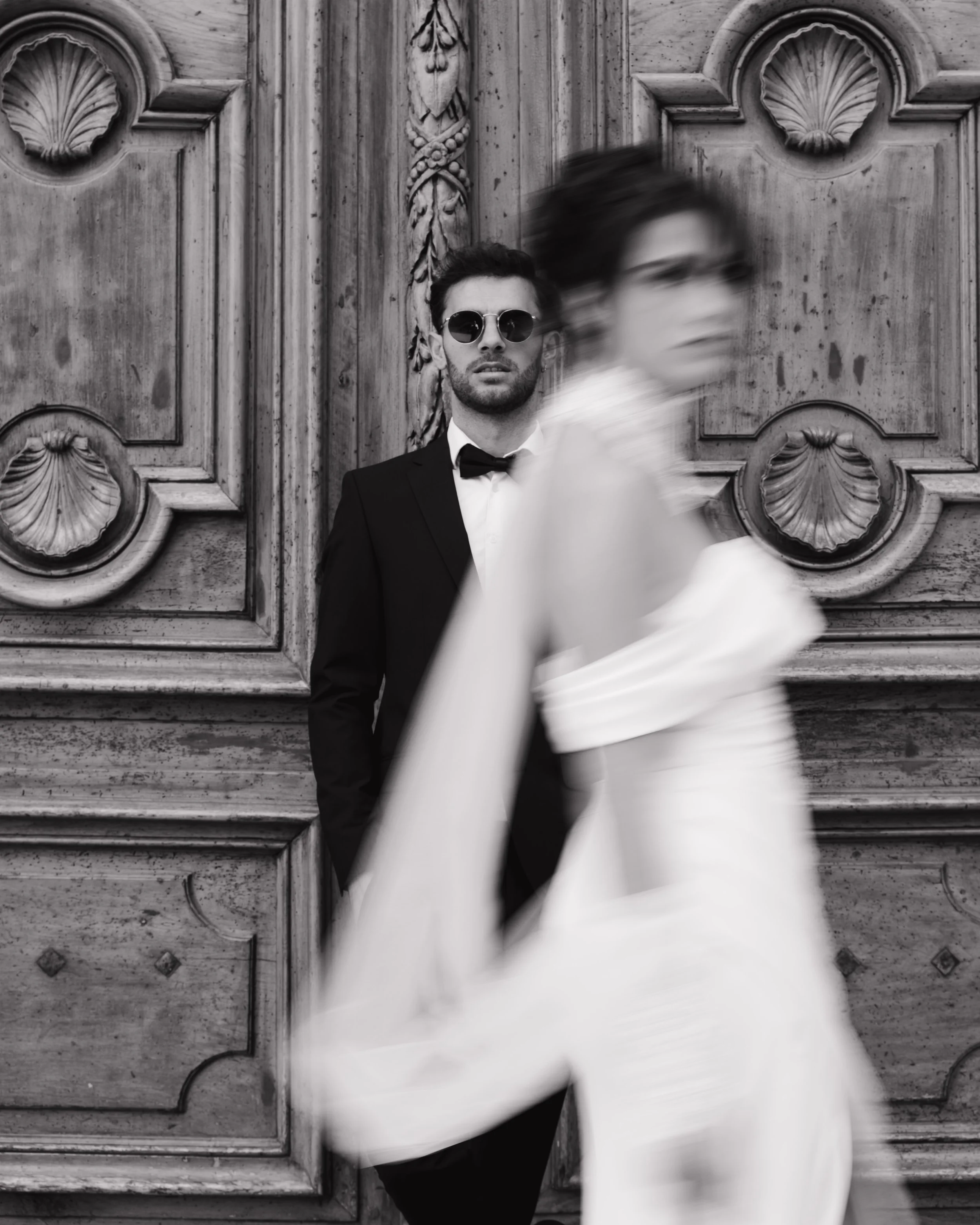 Un homme en smoking avec des lunettes de soleil se tient devant un mur en bois sculpté, tandis qu'une femme en robe blanche passe devant lui en flou à l'hôtel dieu à Lyon à l'intercontinental 