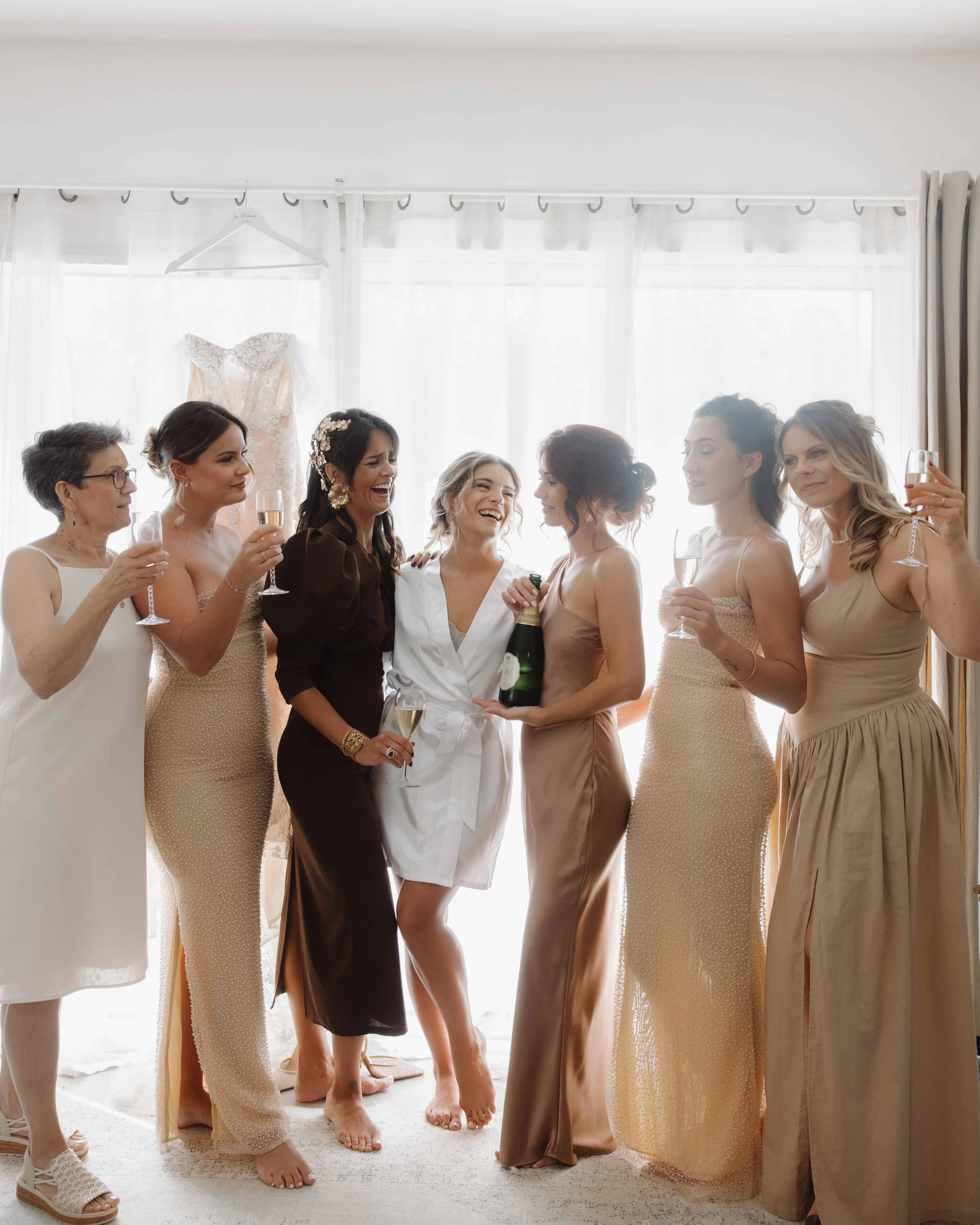 Un groupe de femmes en robe de soirée et une personne en peignoir blanc, tenant une bouteille de champagne, célébrant lors d'une préparation de mariage dans une pièce lumineuse avec une robe de mariée en arrière-plan. Photographe mariage Isère 