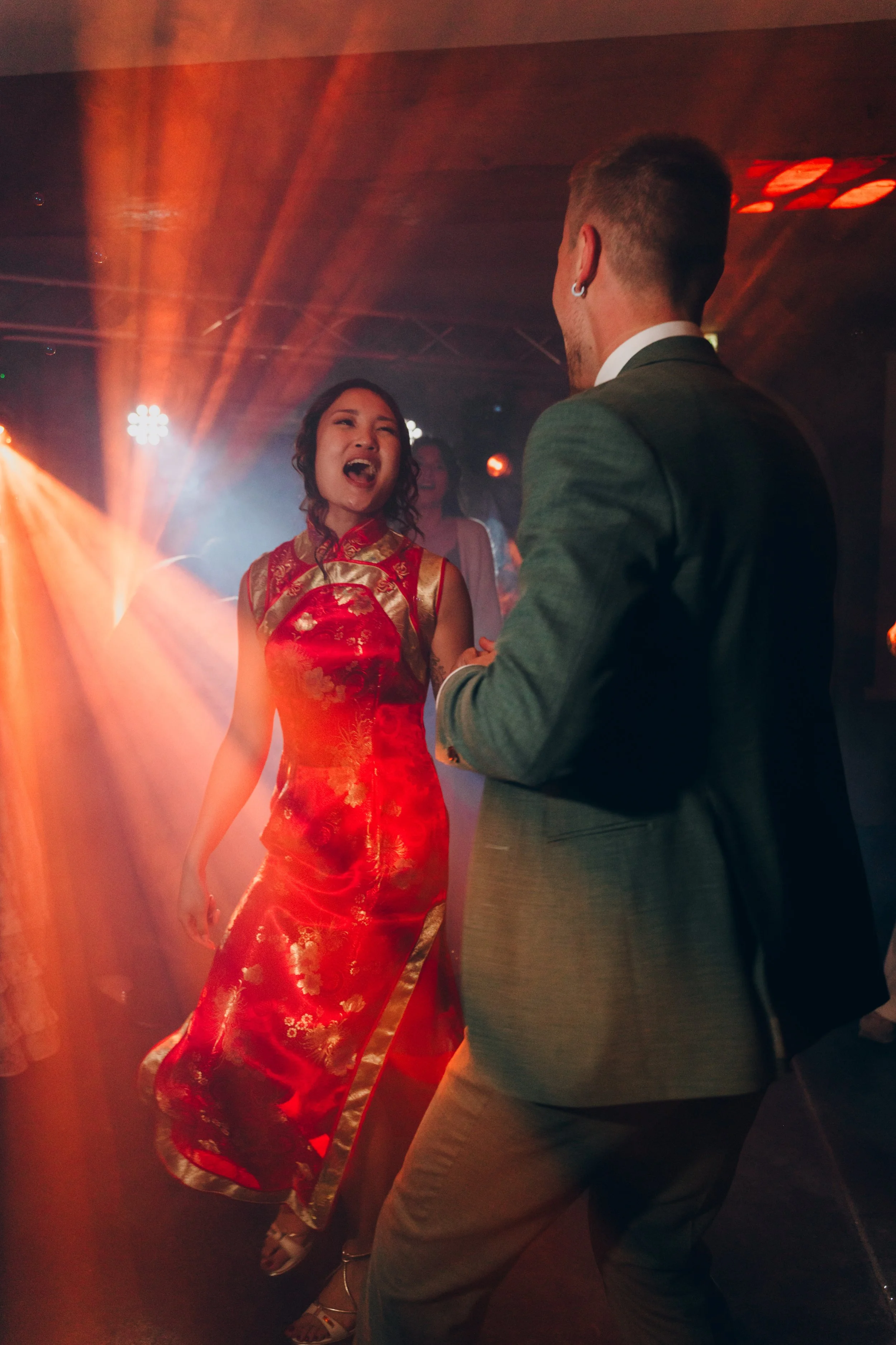Deux personnes dansent avec des lumières de scène colorées en arrière-plan, une femme porte une robe chinoise rouge ornée de motifs dorés, l'autre personne porte un costume gris. Domaine de tourieux à Savigny dans le beaujolais à Lyon. Mariage 