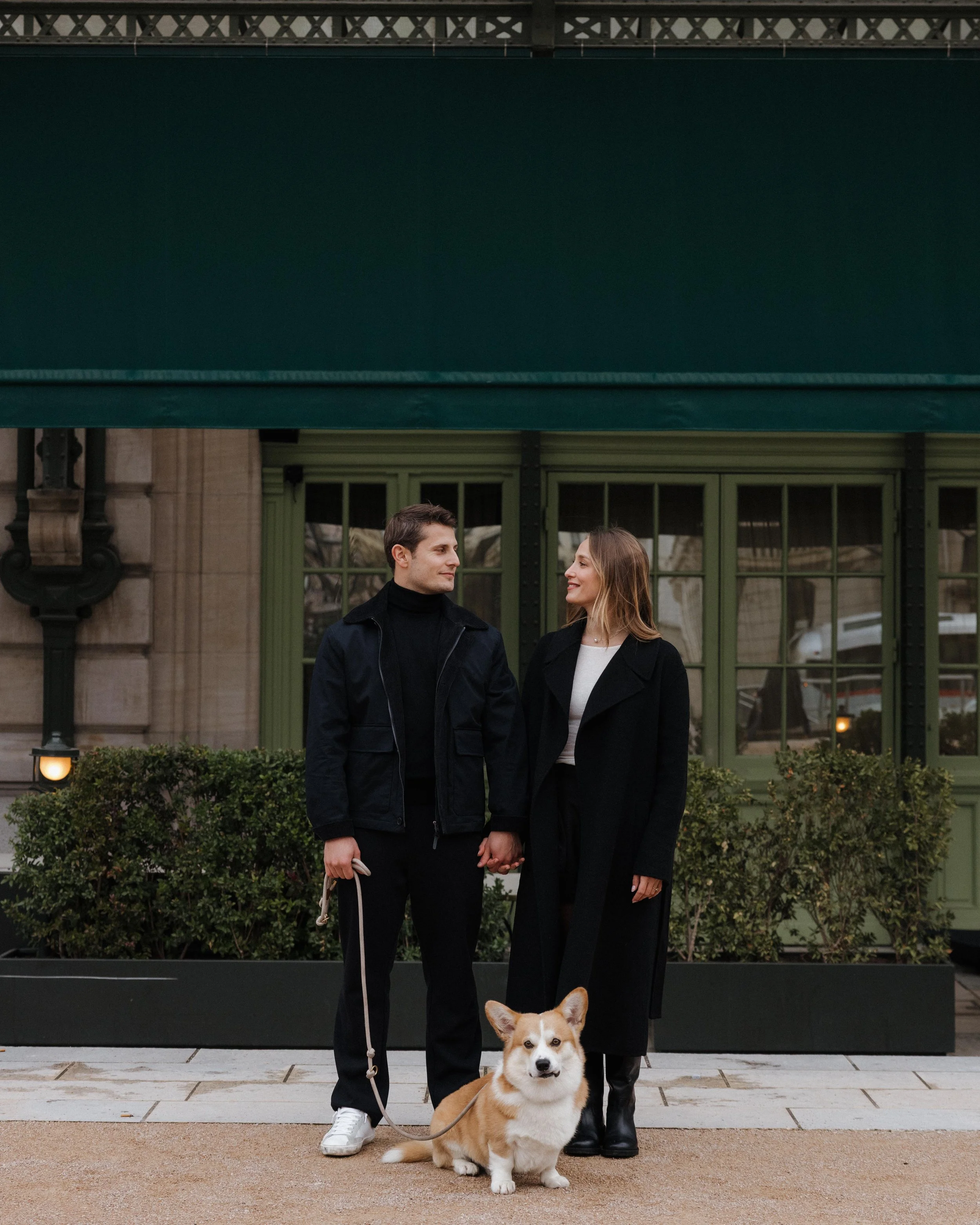 Un couple tenant la main devant un bâtiment avec des fenêtres vertes, accompagné d'un chien corgi beige et blanc, dans un environnement urbain. session engagement couple Lyon 6eme, brotteaux, photographe mariage bordeaux et Lyon et destination weddin