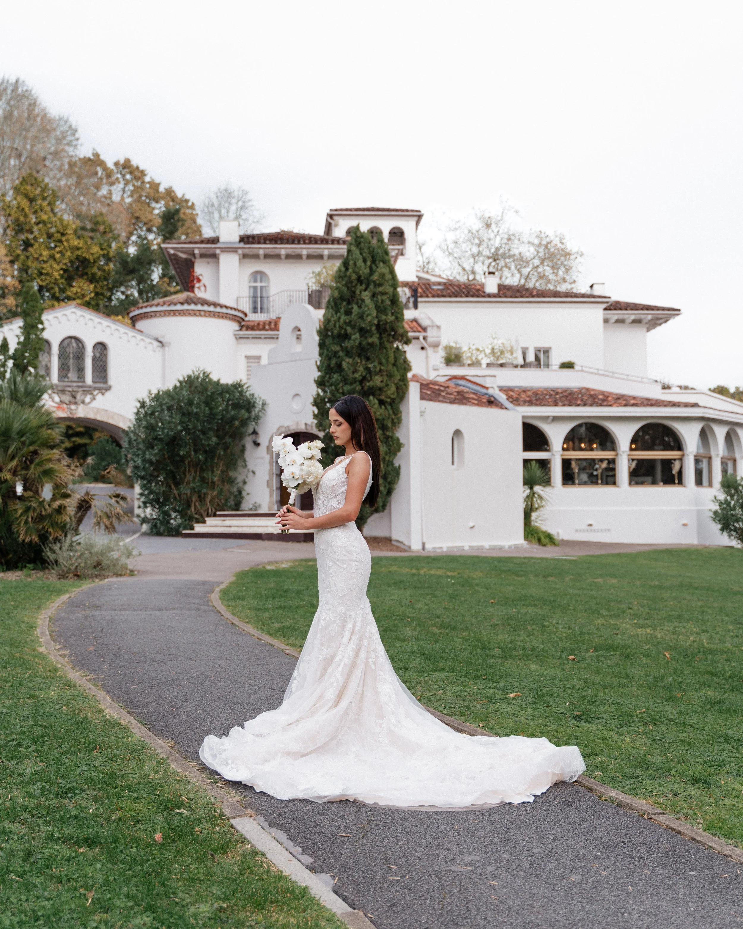 Une femme en robe de mariée blanche tient un bouquet de fleurs blanches devant une grande maison blanche avec un jardin verdoyant.  session femme mariées et robes haut de gamme, brindos, hôtel et lac,  photographe mariage bordeaux et Lyon 
