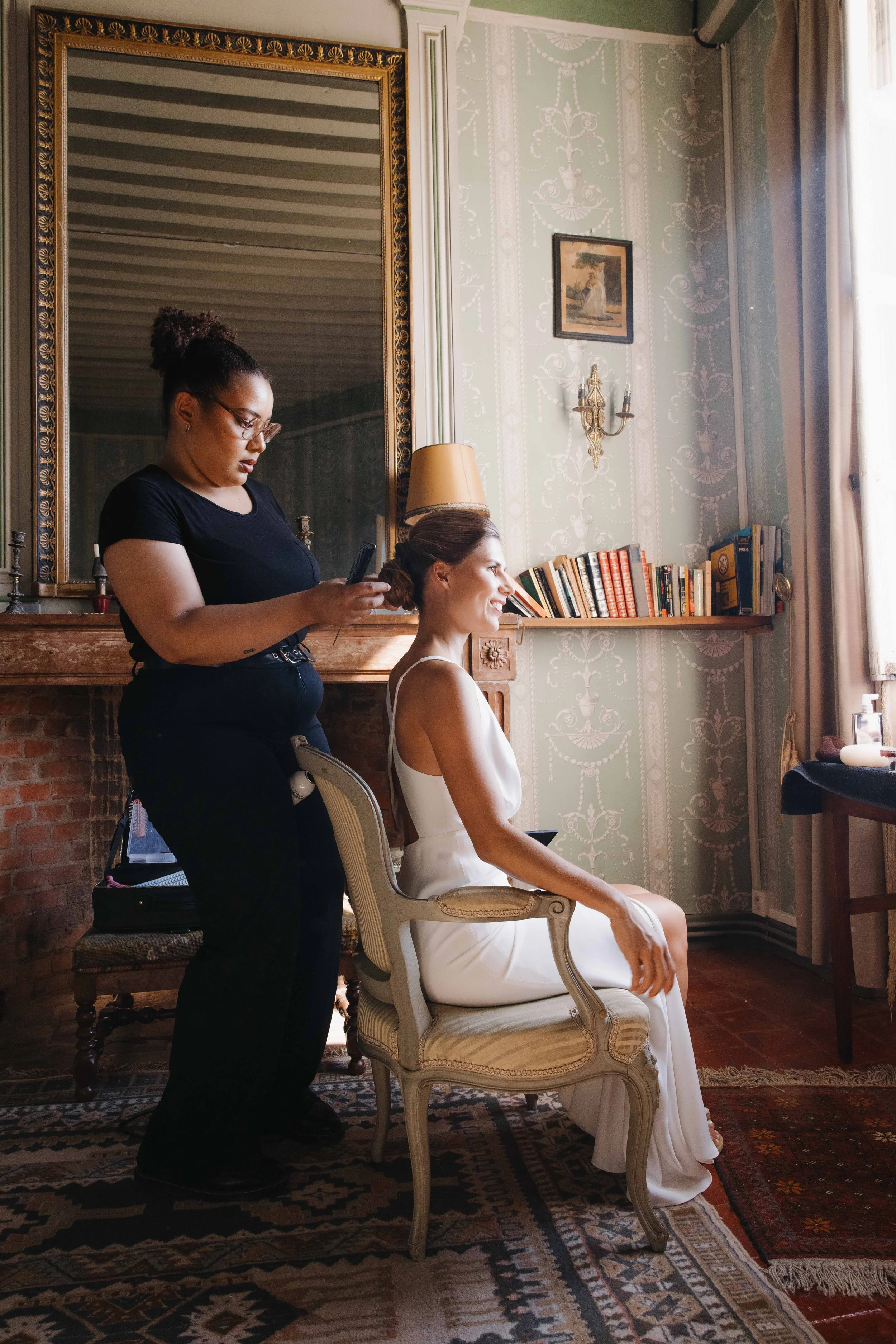Une femme en robe blanche est assise dans une salle ornée d'une grande cheminée en briques et d'une étagère remplie de livres. Une autre femme, en noir, se tient derrière elle, utilisant un téléphone. La scène se déroule près d'une fenêtre laissant e