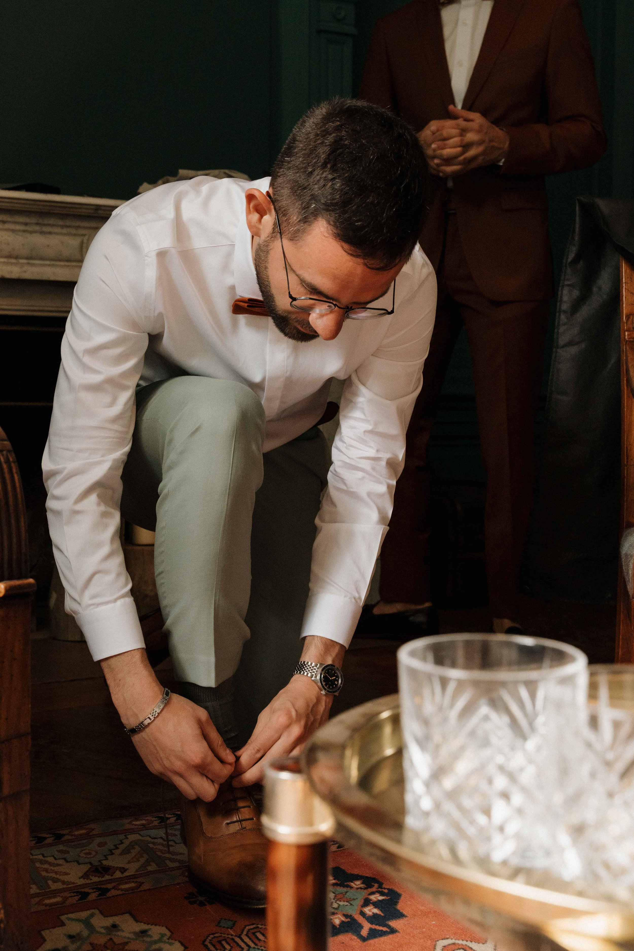 Un homme habillé en blanc avec une cravate marron, en train de lacer ses chaussures en cuir marron, dans une pièce élégante. Domaine de tourieux à Savigny dans le beaujolais à Lyon. Mariage cérémonie laïque, photographe Lyon haut de gamme, duo 