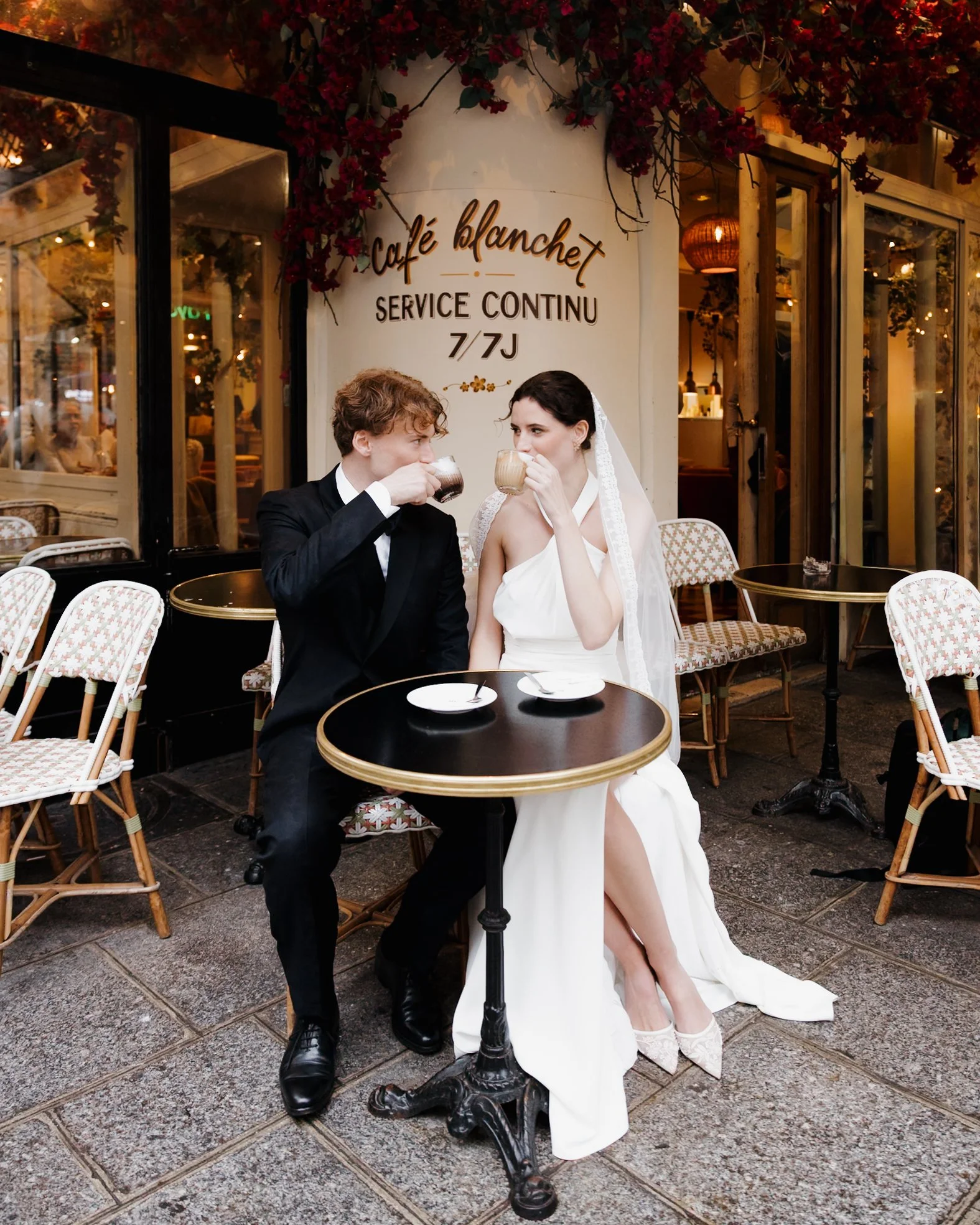 Un couple habillé en mariage, assis à une table en terrasse de café, buvant un café ensemble devant un restaurant appelé 'Café Blanchet'. Session engagement café parisien, photographe mariage paris haut de gamme, destination wedding 
