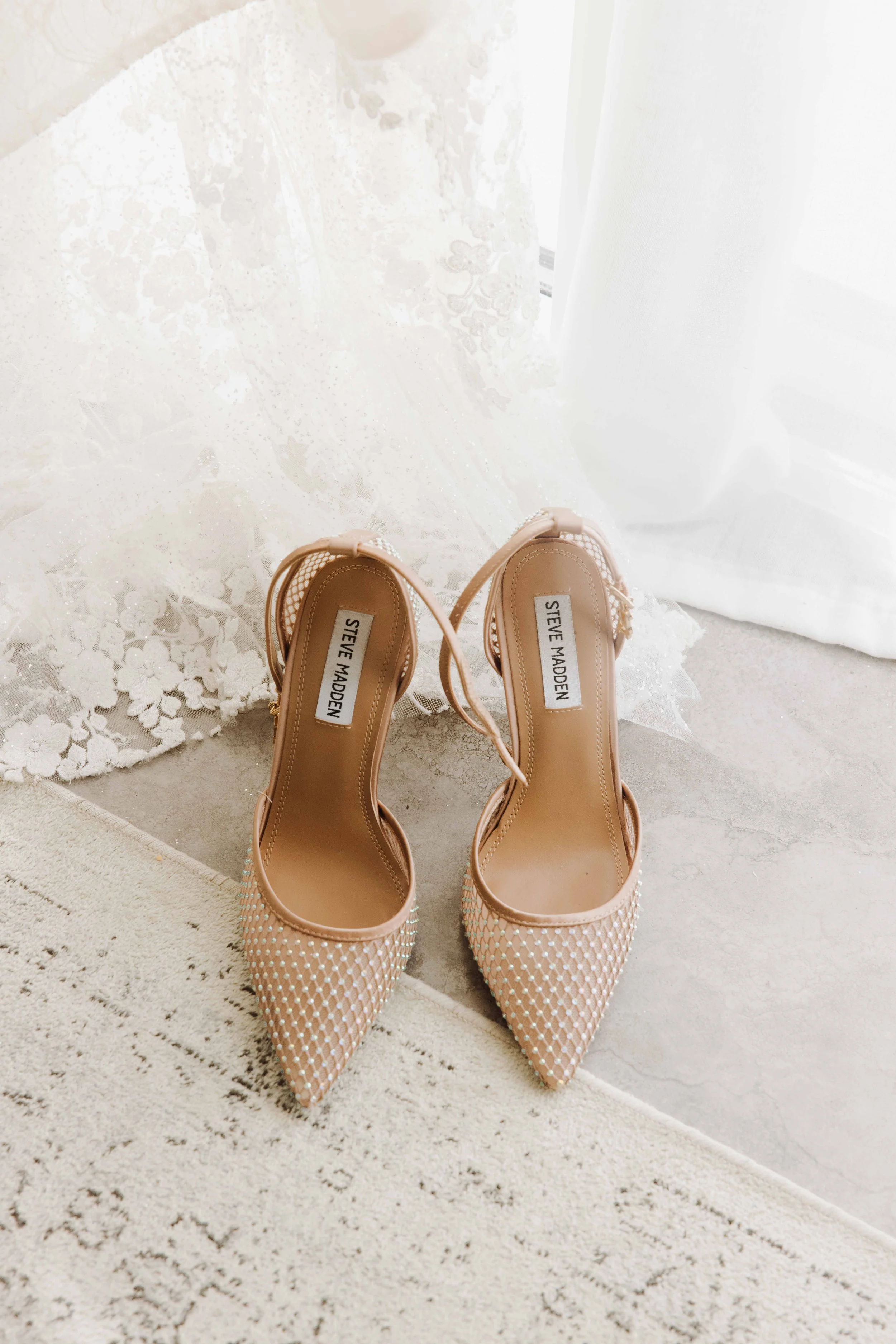 Chaussures à talons blancs avec détails perlés, de marque Steve Madden, posées sur un tapis beige près d'une fenêtre avec rideau blanc. Photographe mariage Isère, ferme du rocher, cérémonie civil et laïque. Bordeaux et Lyon et destination wedding.