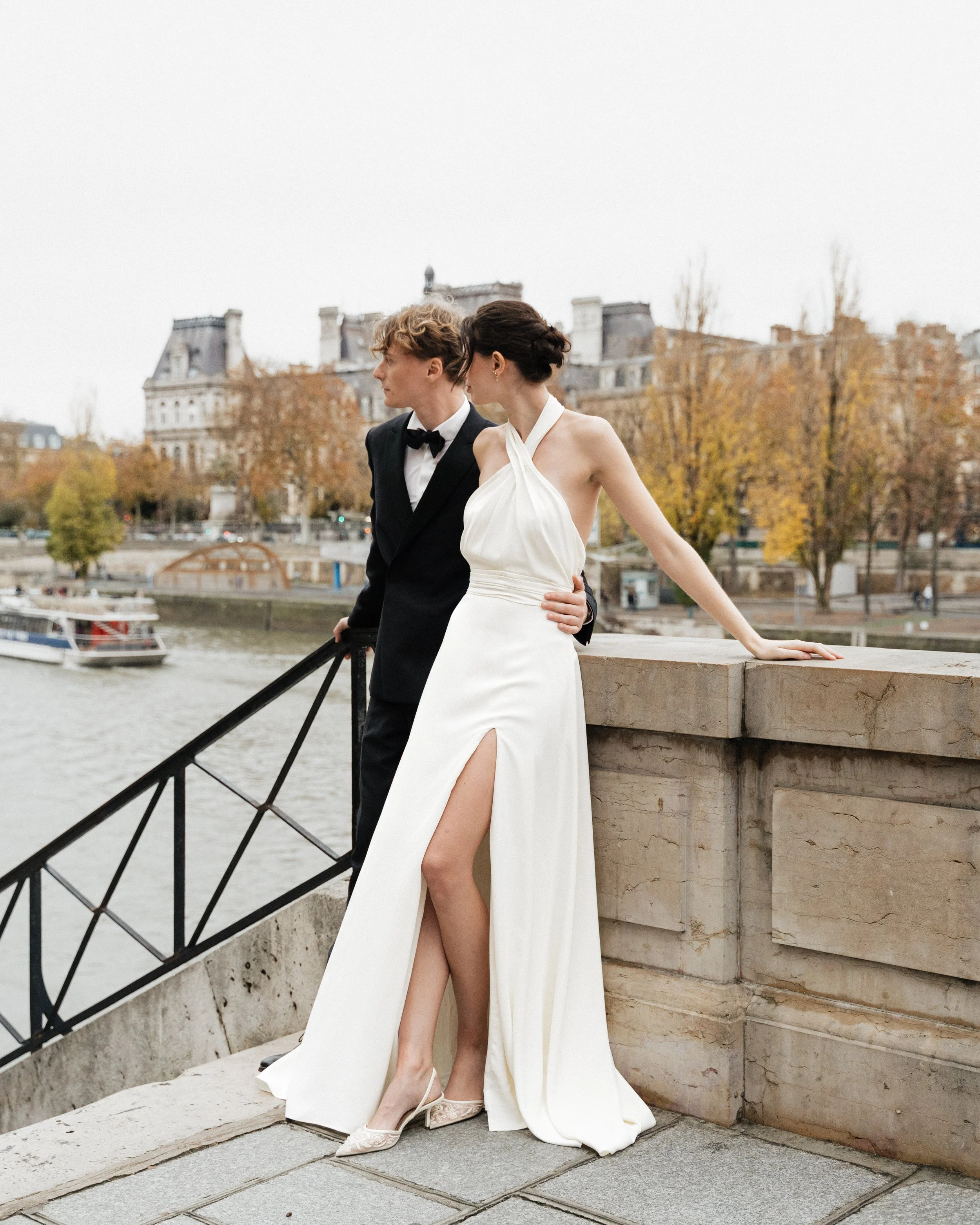 Un couple en vêtements de mariage se tenant par la taille sur un pont à Paris, avec la Seine et des bâtiments historiques en arrière-plan. Session engagement paris, Louvre, mariage haut de gamme, destination wedding. Robe de mariées, madame a dit oui