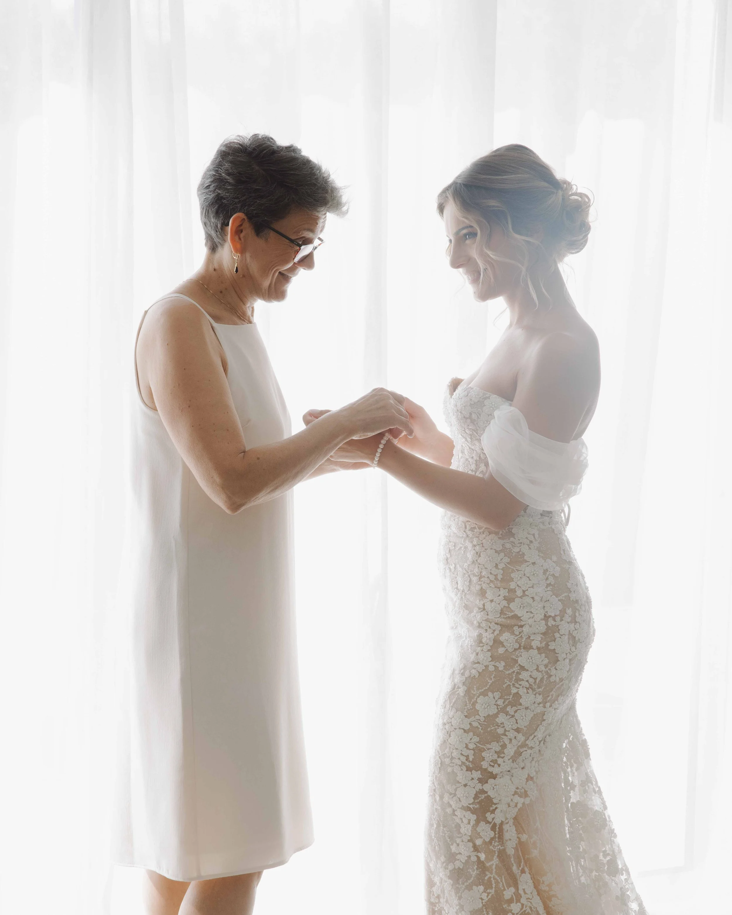 Une jeune mariée en robe de mariage blanche sourit pendant qu'une femme plus âgée, probablement sa mère, lui reprend la main et lui ajuste un bracelet, devant une fenêtre lumineuse.