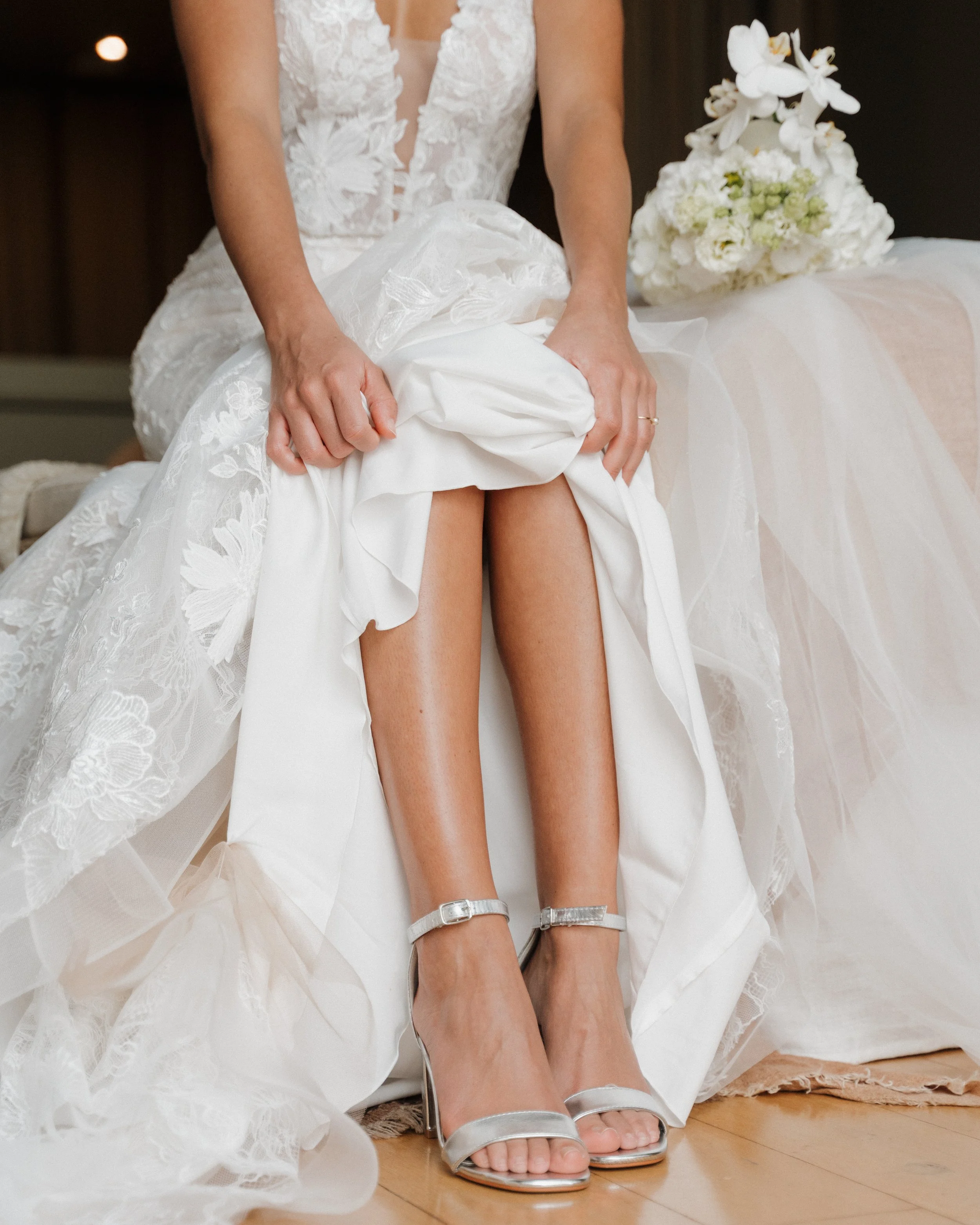 Une femme en robe blanche de mariage retire ou ajuste sa jupe pour révéler ses jambes et ses chaussures à talons argentées avec une bride. Un bouquet de fleurs blanches est visible à l'arrière-plan.  session femme mariées et robes haut de gamme 