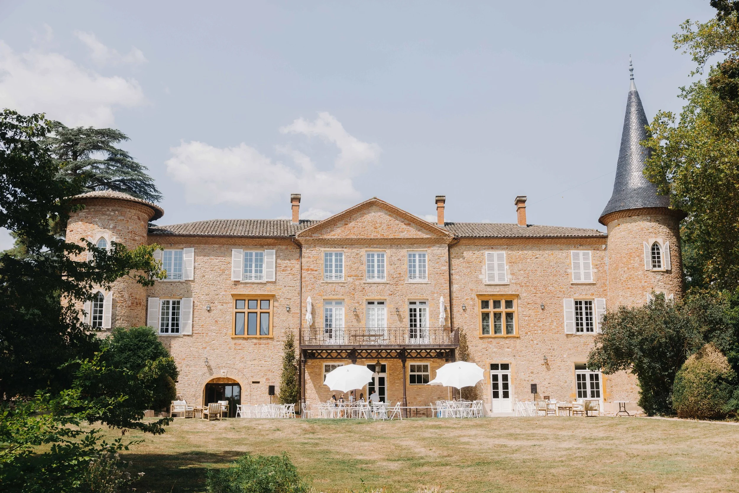 Château en pierre avec tours rondes et tourelles, fenêtres à volets blancs, terrasse avec parasols et mobilier d'extérieur, arbres et herbe devant, ciel partiellement nuageux. Mariage chateau de champ renard, Blacé, beaujolais, Lyon, préparatifs