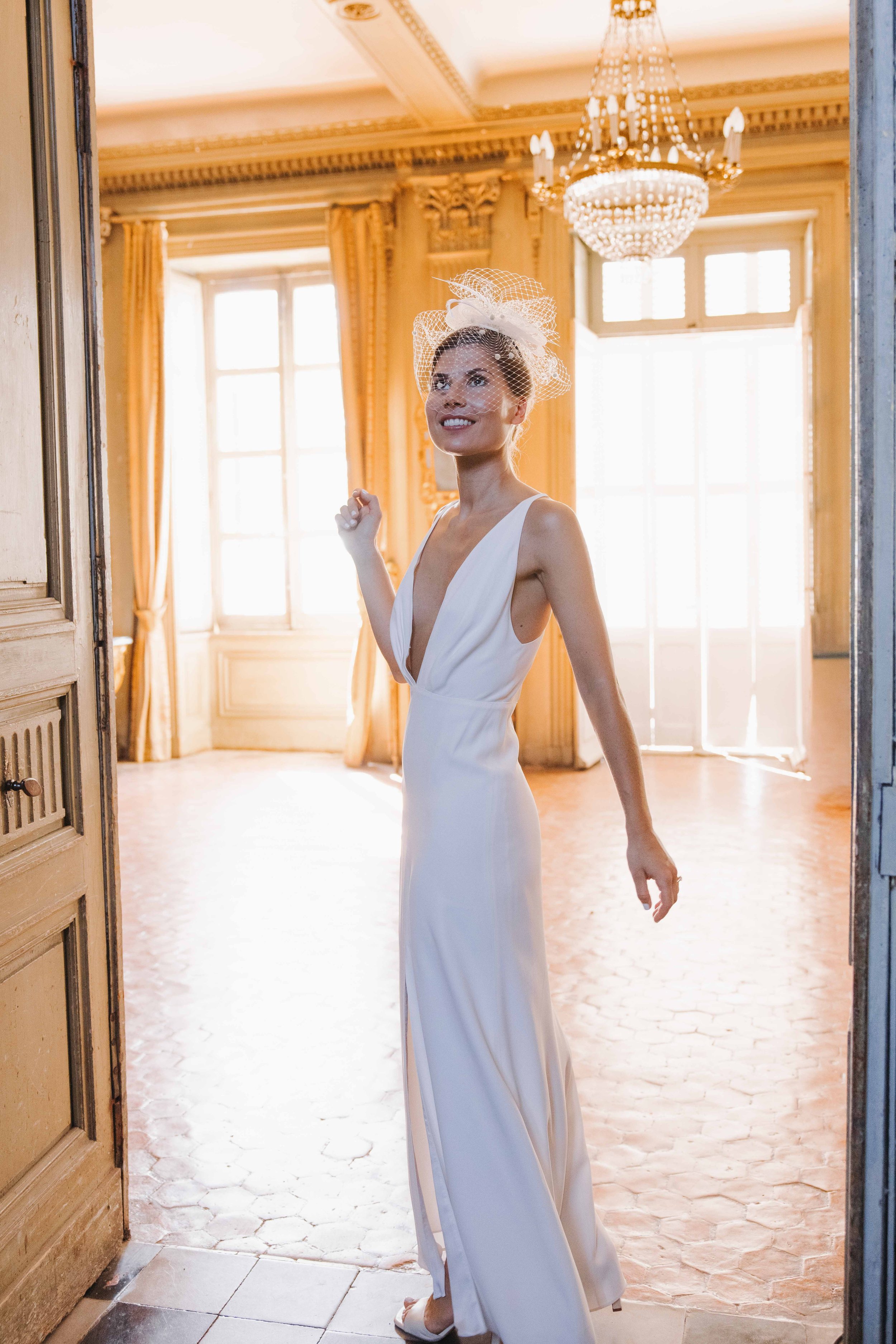Une femme en robe de mariée blanche, souriante, portant un voile avec un fascinator, dans une pièce élégante avec de grandes fenêtres et un chandelier au plafond. Mariage au chateau de lachal dans le rhone alpes à côté de Lyon. Cérémonie civil 